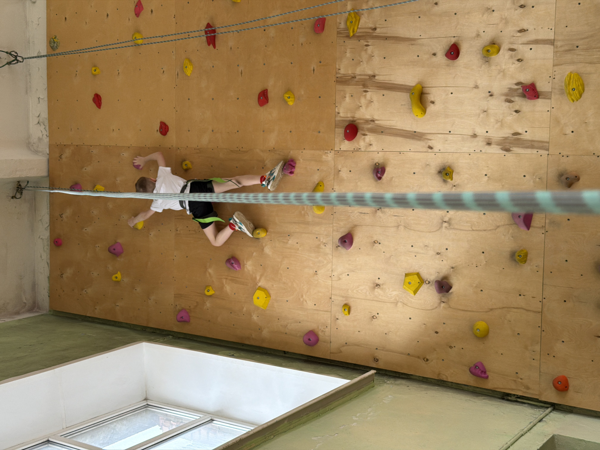 A child is climbing an indoor rock climbing wall. The wall is made of wood and has various colored holds attached to it. The child is wearing a white shirt, black shorts, and climbing shoes. They are secured with a harness and a rope that runs horizontally across the wall. The location appears to be in Izhevsk, Russia, based on the presence of unique structural features and architectural styles known in the area. A window is visible in the lower left corner, and the flooring is green.
