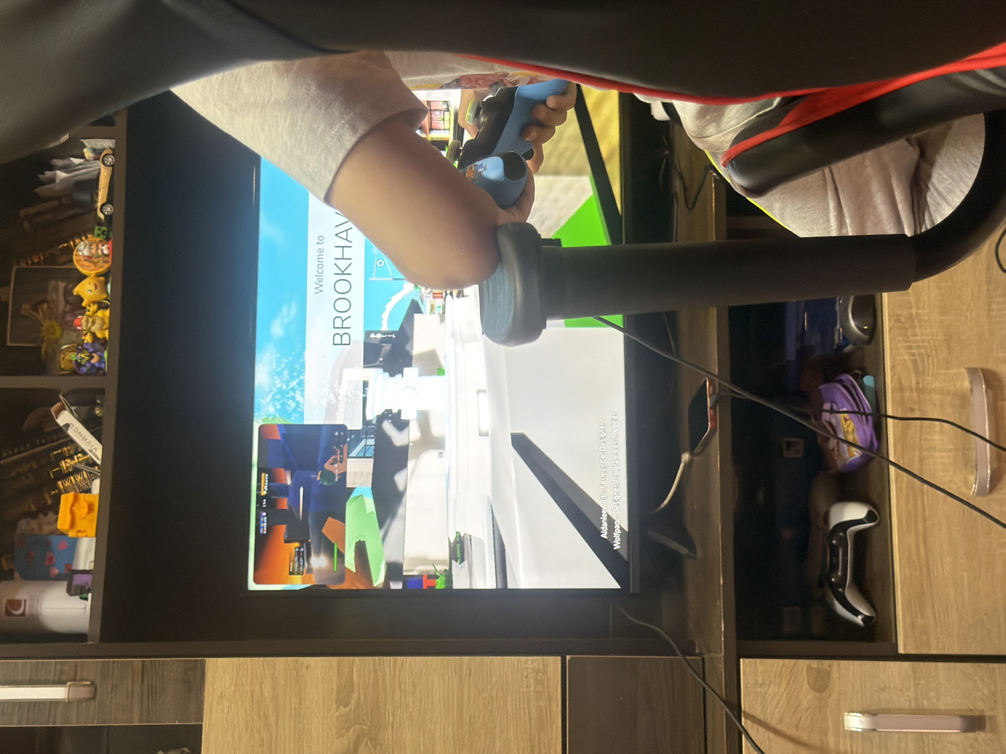 An overhead view captures a person, likely a child or young adult, from the chest down, engaged in playing a video game. The person is seated in a gaming chair with red and black accents, and their arm rests on a black armrest. Both hands are holding a light blue gaming controller, indicating active gameplay.
The focal point is a large television screen displaying a video game. The screen shows the text "Welcome to BROOKHAV," which is likely the title screen for the popular Roblox game "Brookhaven." Below the title, credits are visible, listing "Aldanleewalt - Former Co-Designer" and "Wolfpaq - Designer, Programmer, Modeler." The game graphics are stylized and blocky, characteristic of Roblox.
The setting appears to be an indoor room, possibly a gaming corner or bedroom. To the left of the screen, a wooden shelving unit holds various items, including a skateboard, small figurines (such as Pokémon characters), books, a blue and pink patterned mug, and a white object resembling a toy plane. An item on one shelf is labeled "VIVANCO." To the right of the screen, another wooden storage unit is visible, with a purple bag and a white and black gaming controller tucked into an open compartment. Several cables are visible around the setup, indicating connected electronic devices. The overall impression is a cozy, personal gaming space.