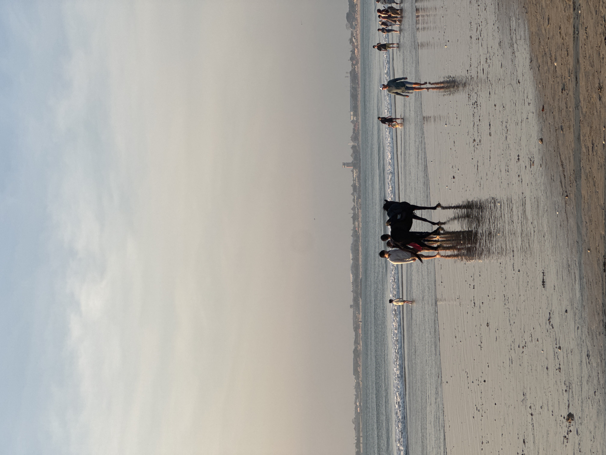 The image captures a tranquil scene on a beach in Kuta Selatan, Indonesia, during what appears to be late afternoon or early evening. The sky is a soft gradient of pale blue and dusty orange, with wispy clouds scattered across the horizon. The sea is calm, with gentle waves lapping at the shore, reflecting the muted light of the sky. The wet sand of the beach is speckled with small stones and debris, and it gleams with the reflections of the sky and the subjects present.
In the foreground, a dark horse stands partially submerged in the shallow water, with two individuals standing beside it. One person, dressed in a light-colored shirt, is facing away from the camera, while another person, wearing darker clothing, is also in close proximity to the horse. Further down the beach, a group of people are visible walking along the shore. A lone individual in a white shirt stands in the shallow water, facing the sea. To the right, a cluster of people are walking together, some in casual attire, suggesting they are enjoying a leisurely stroll. In the distance, a faint silhouette of buildings and structures can be seen along the coastline, hinting at the presence of a town or resort. The overall atmosphere is peaceful and serene, with the natural beauty of the beach and the subtle human activity creating a harmonious composition.