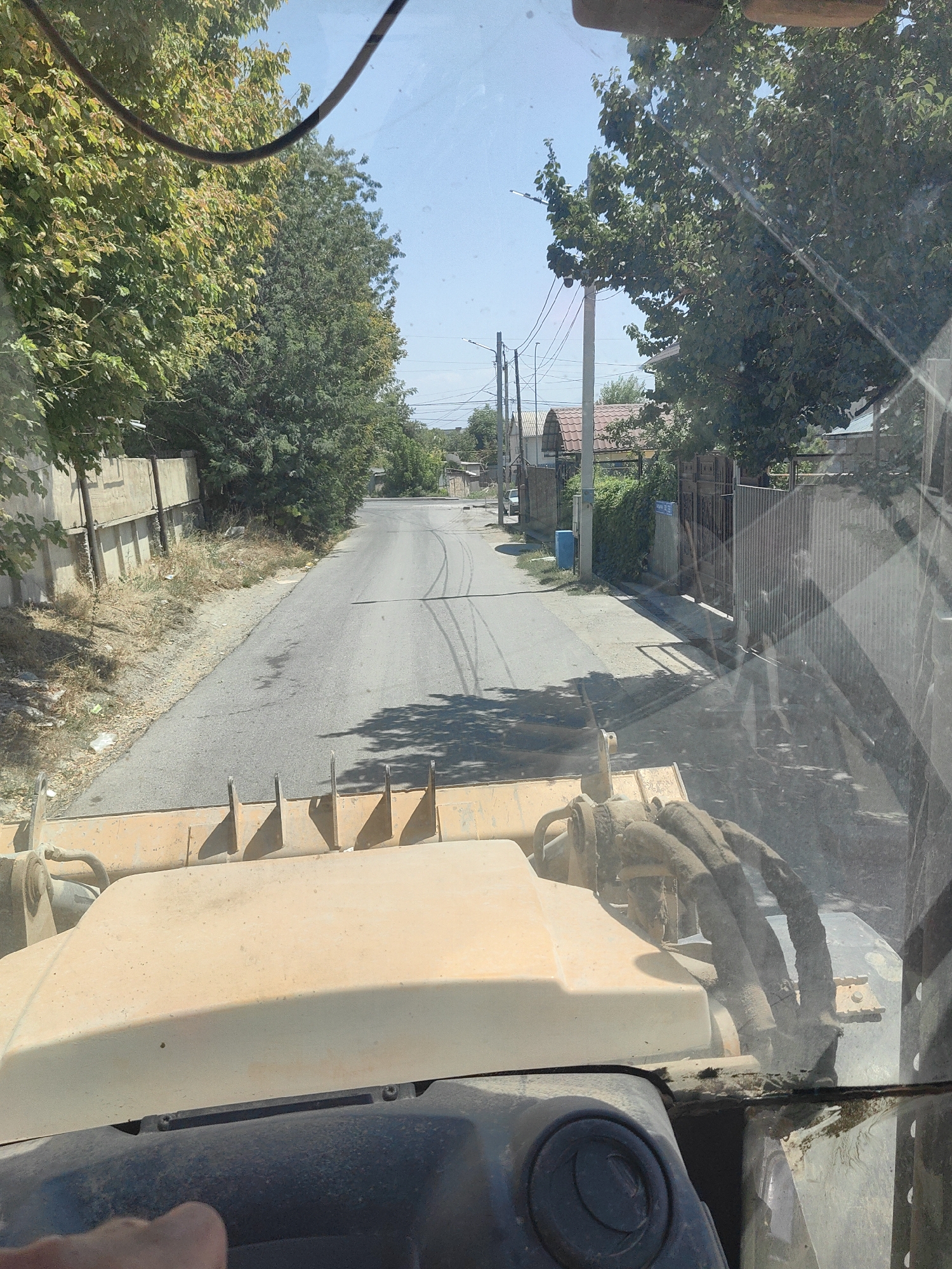The image is taken from the driver's perspective inside a vehicle, likely a construction vehicle due to the large yellow front loader visible at the bottom. The vehicle is on a paved road in what appears to be a residential area. Trees line both sides of the road, with some houses and fences visible in the background. The sky is clear and blue. The location is identified as the city of Shymkent, Kazakhstan. The scene depicts a sunny day with clear visibility. The road shows tire marks, suggesting recent vehicle activity. There are no people visible in the immediate foreground, and the focus is on the road and the surrounding environment.