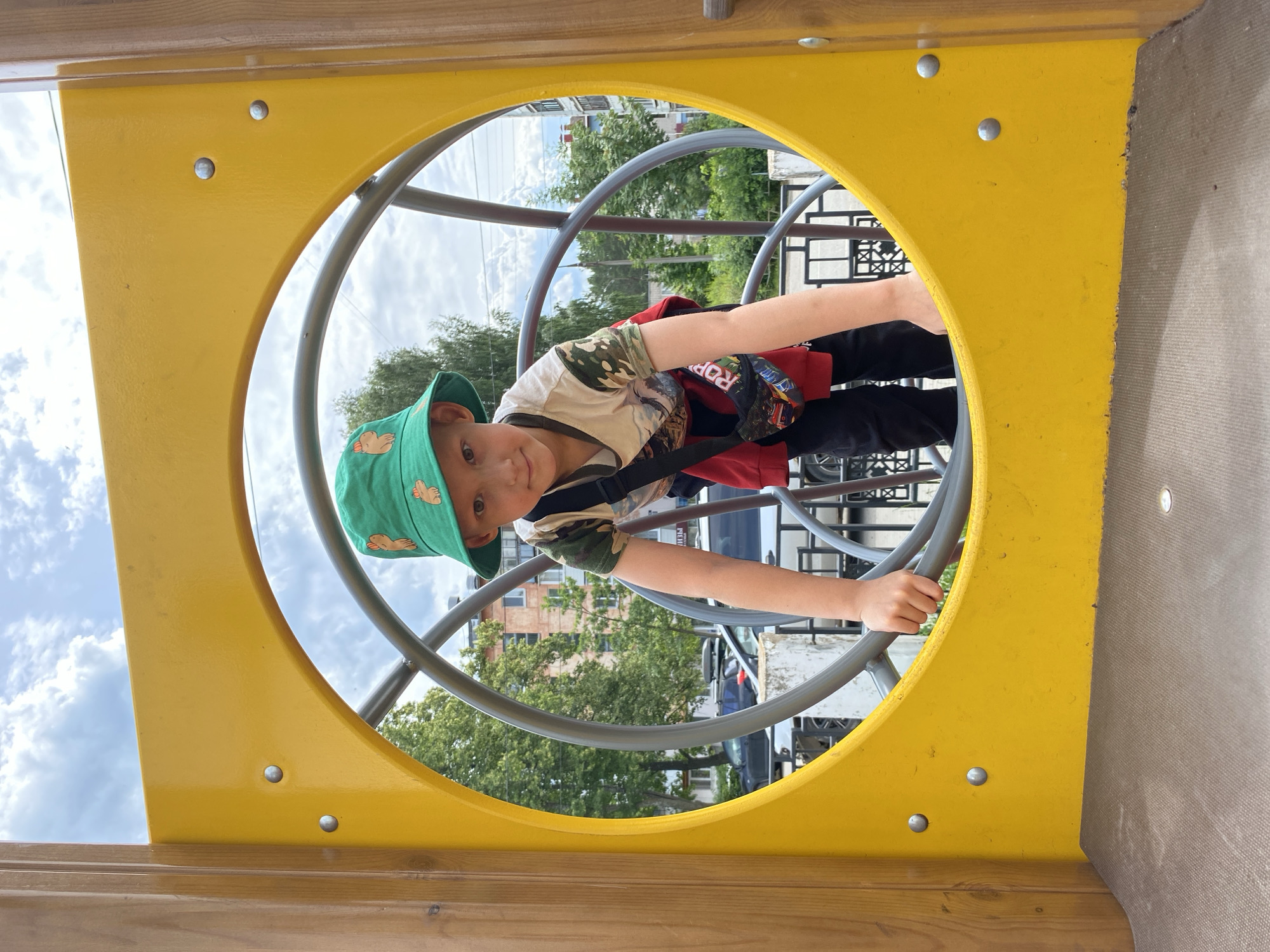 An outdoor shot captures a young boy on a playground in Chaikovsky, Russia. He is wearing a green bucket hat with a repeated cartoon animal print, a t-shirt with a camouflage pattern on the sleeves and a graphic design on the front, black pants, and a red hooded jacket tied around his waist. A small black fanny pack with a colorful car graphic is worn across his chest. The boy is positioned within a large, circular opening of a yellow playground structure made of wood and metal. He is holding onto the metal bars of the structure with both hands, appearing to be climbing or in the process of moving. His body is leaning forward, and he is looking directly at the camera. Behind the boy, through the circular opening, a cloudy sky is visible, along with trees and buildings, indicating an urban or suburban setting. The ground beneath the playground structure is covered with a textured, gray material.