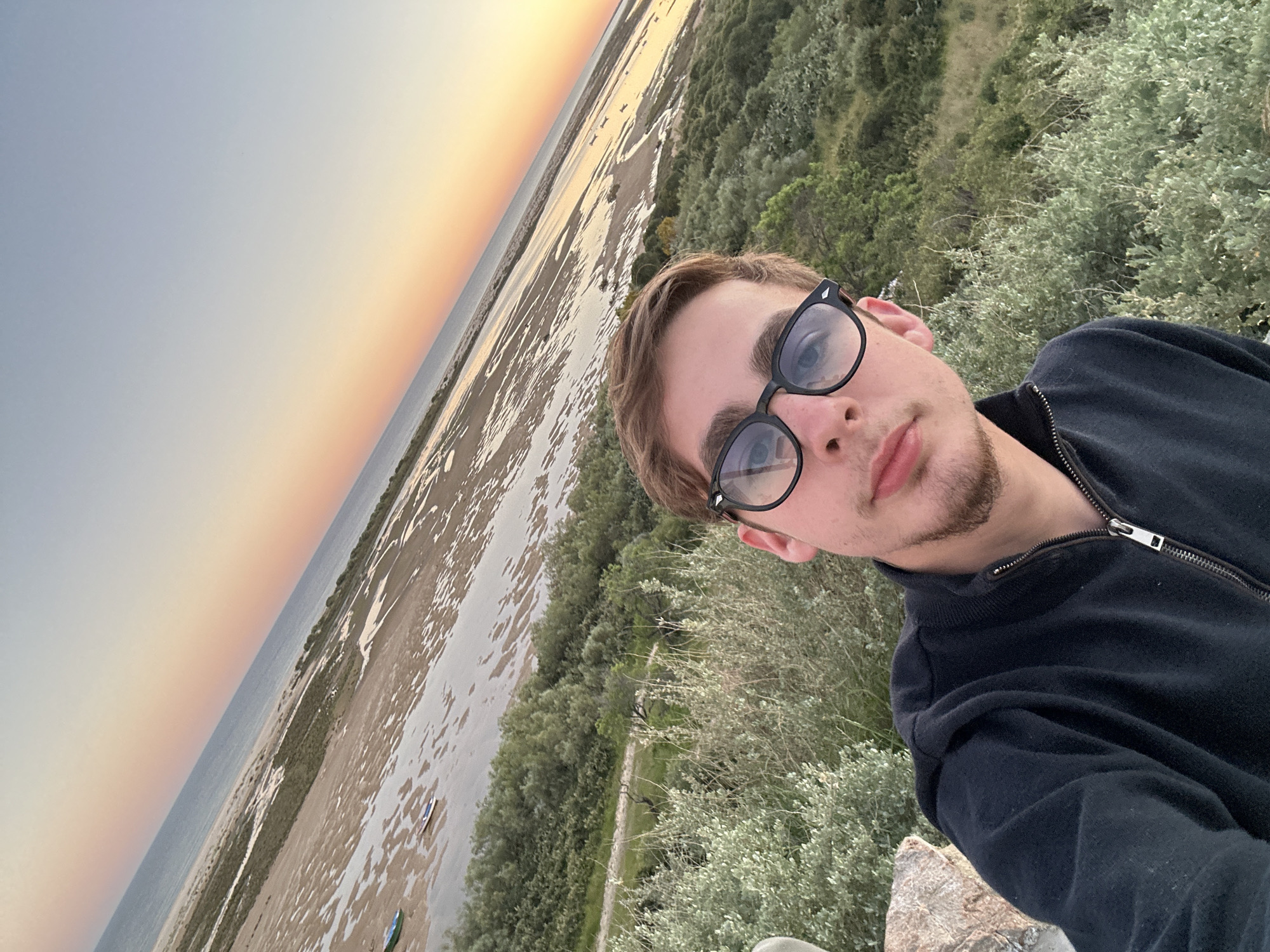 The image is a selfie taken outdoors, possibly in Vila Real de Santo António, Portugal. A young man with short brown hair, a light beard, and wearing black-rimmed glasses is the central figure. He is wearing a dark jacket that is partially zipped up. He appears to be standing on a slightly elevated position, possibly a rocky outcrop, surrounded by low-lying green vegetation. In the background, there is a view of a flat, sandy landscape interspersed with bodies of water, likely a coastal area or river delta. The sky in the distance shows a gradient of colors, suggesting either sunrise or sunset. The overall composition is oriented at an angle, giving the impression of a dynamic, informal snapshot.