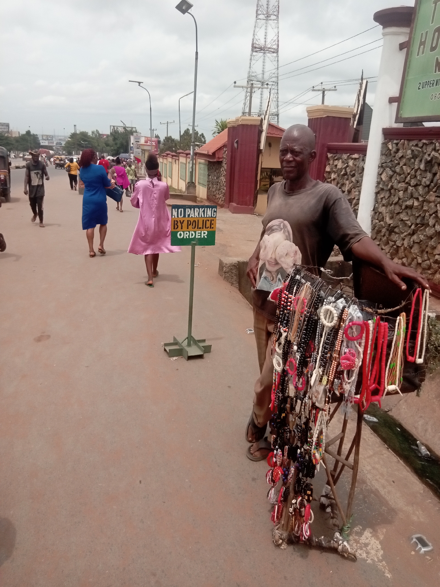 A man stands on the side of a road in Onitsha, Nigeria, beside a display of necklaces and bracelets on a small stand. The road is a light brown color. The man is dark-skinned, with a shaved head, and wears a faded t-shirt with a graphic, brown trousers, and sandals. Behind him is a building with a stone facade and a sign that says "HO...N". A "NO PARKING BY POLICE ORDER" sign stands between the man and a group of people walking away from the camera down the street. In the background, there is a communications tower, street lights, and more buildings, all under a cloudy sky.