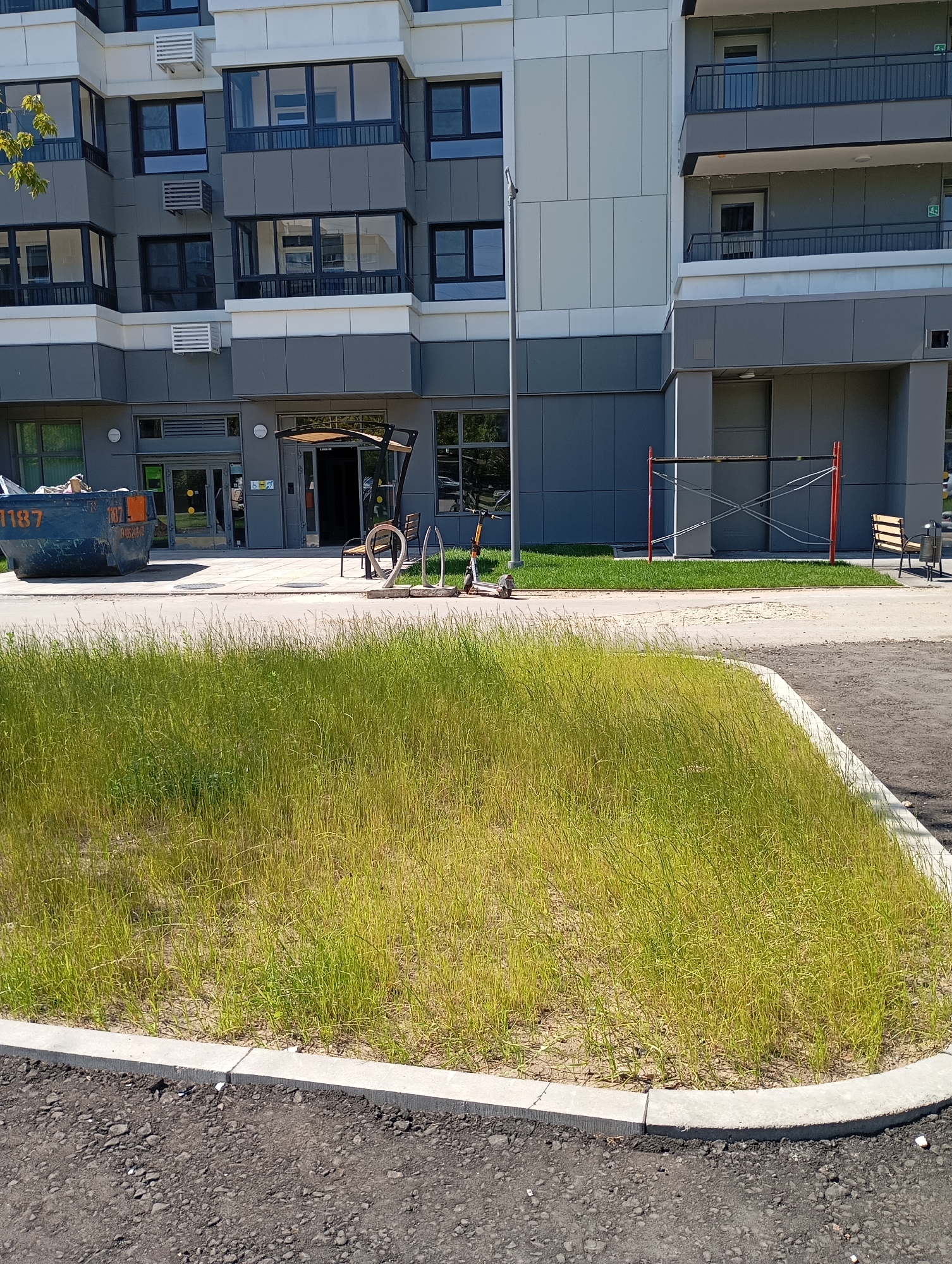 The image depicts the exterior of a modern residential building in Moscow, Russia, under a clear sky. In the foreground, a strip of tall, dry grass is bordered by asphalt pavement and a concrete curb. Behind the grass, a paved area runs along the building's facade. To the left, a large, dark blue dumpster with the number "1187" visible on its side is positioned near the entrance. The building features multiple floors with a grey and white facade, numerous windows, and balconies. On the paved area near the entrance, a covered outdoor seating area with a bench is situated. A bicycle rack holds two bicycles, and an electric scooter is parked to the right of the bicycles. Scaffolding with red poles and crossbars is erected against the building to the right of the covered seating area. The overall scene suggests an urban residential environment, possibly under construction or recently completed, given the presence of the dumpster and scaffolding.
