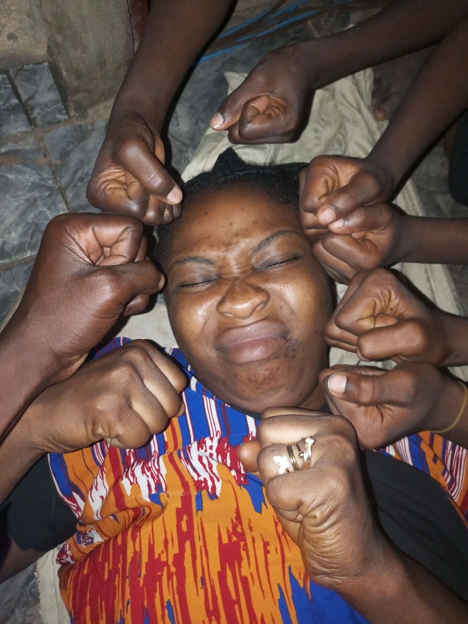 A woman is lying on her back with her eyes closed and her face contorted in distress. Several hands are reaching towards her, some appearing to be in a fist. She is wearing a colorful striped shirt. The scene is indoors, with a textured gray floor and what appears to be a dirt or concrete wall in the background. The lighting is dim. This image was taken in Tempure, Nigeria.