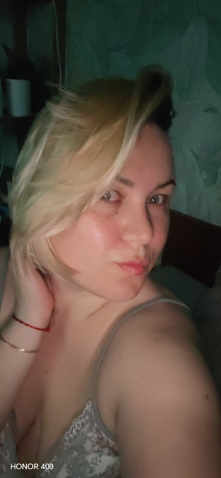 The media file features a close-up selfie of a woman taken indoors in Noviy, Russia. The woman has short, blonde hair, fair skin, and light-colored eyes. She is looking directly at the camera with a slightly pouty expression, and her left hand is raised towards her neck and hair. She is wearing a light-colored camisole with visible lace detailing on the strap and bust. On her left wrist, she has a red string bracelet and a thin gold chain bracelet. The background is dimly lit and appears to be an indoor room, possibly a bedroom, with a textured or patterned wall. In the upper left portion of the image, vague outlines of objects, including what resembles a bottle and a white cylindrical item, are visible on a shelf or surface. The low lighting suggests the photo was taken in the evening or at night. The text "HONOR 400" is visible as a watermark in the bottom left corner of the image.