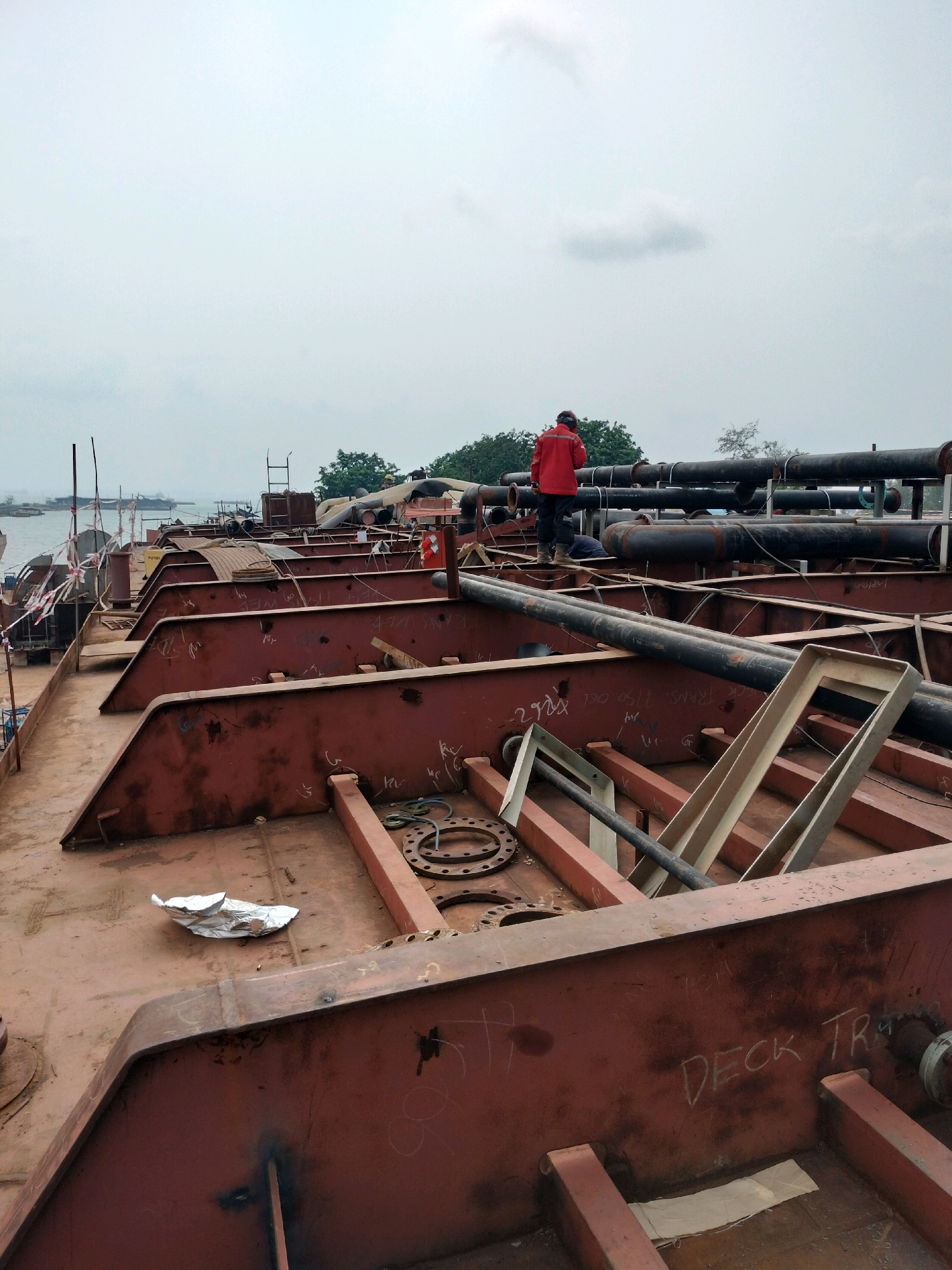 A person wearing a red jacket and a hard hat stands on a large, rust-colored metal structure, possibly a ship or barge under construction or repair, in Batam, Indonesia. The structure has a complex network of large pipes running across it. In the foreground, there are angled metal beams and a large metal flange. In the background, there is a body of water with a few boats and lush green trees on the horizon. The sky is overcast. The overall scene suggests an industrial setting, likely a shipyard or port facility.