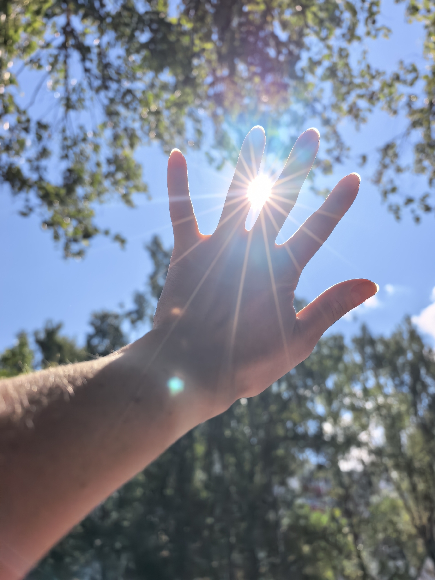 The image shows a person's hand held up towards the sun, with the fingers spread. The sun is shining brightly through the fingers, creating a starburst effect. The background is a blue sky with some blurry green foliage. There is no indication of the city of Sterlitamak, Russia, or any specific location. The image is a close-up shot of the hand and the sun.