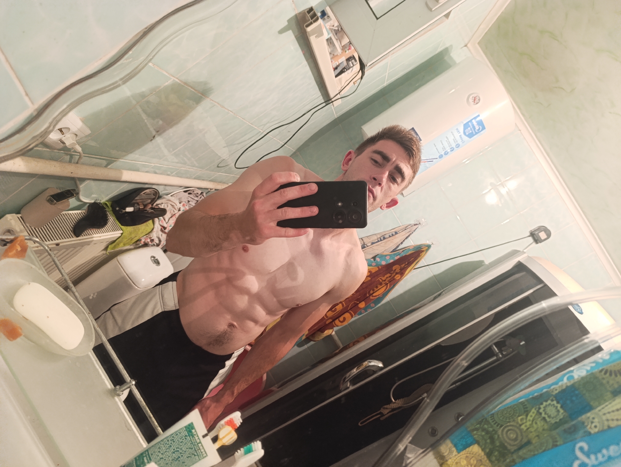 The media file is a selfie taken in a bathroom. The subject is a shirtless man with well-defined abdominal muscles, holding a smartphone to take the picture. He is wearing black shorts. The bathroom appears to be tiled with light blue and green tiles. Reflecting in the mirror, we can see a toilet, a sink with a bar of soap, a toothbrush and toothpaste, a water heater, a radiator, and some towels. The lighting suggests it is likely daytime, as there are no strong artificial light sources visible besides what is reflecting from the room itself. The overall atmosphere is casual and private, typical of a personal photo taken in a bathroom. No discernible text is visible in the image.