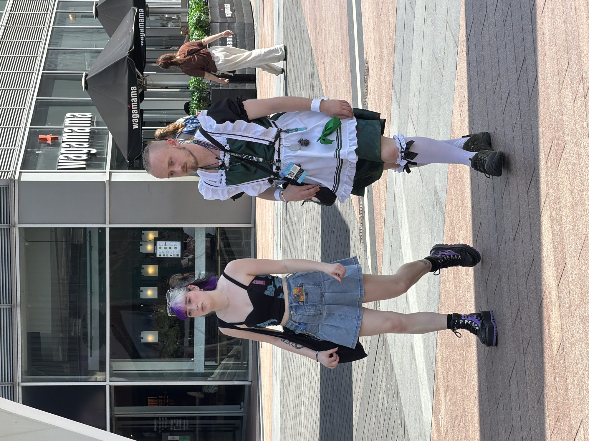 The image captures two individuals standing on a paved area in what appears to be a public space. Behind them is a building featuring the "wagamama" restaurant logo. One person is dressed in a green and white maid costume complete with stockings and black shoes. The other person is wearing a black top, denim skirt, and black platform boots with purple accents. The area appears to be in London, United Kingdom, based on the restaurant name and architectural style. The people are standing still, facing the camera.