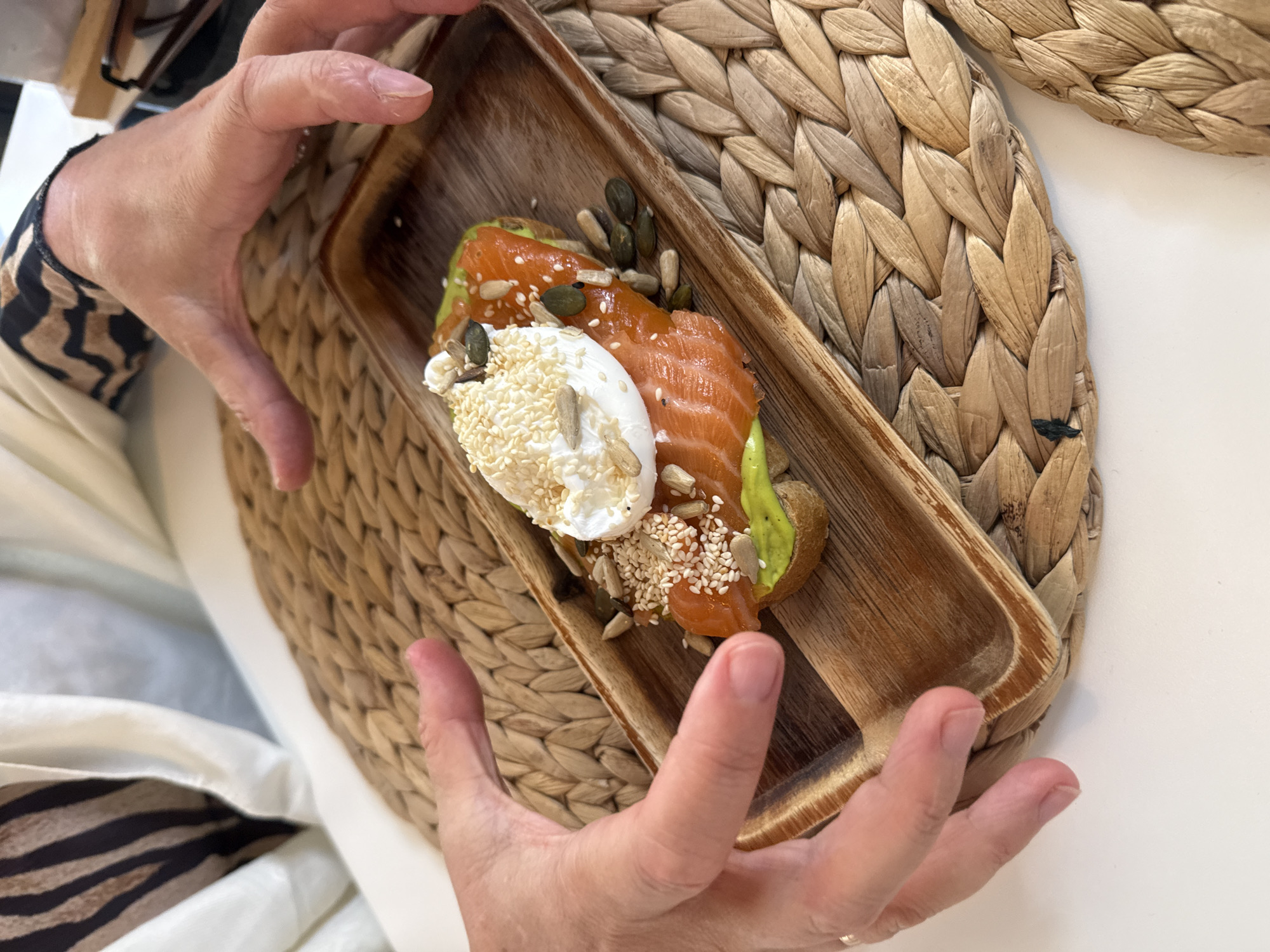 A person's hands are holding a wooden tray containing an open-faced sandwich. The sandwich is topped with sliced avocado, smoked salmon, a poached egg, and various seeds including sesame, sunflower, and pumpkin. The tray rests on a woven placemat on a white surface. The background shows a person wearing a white and brown patterned top. The scene appears to be indoors.
