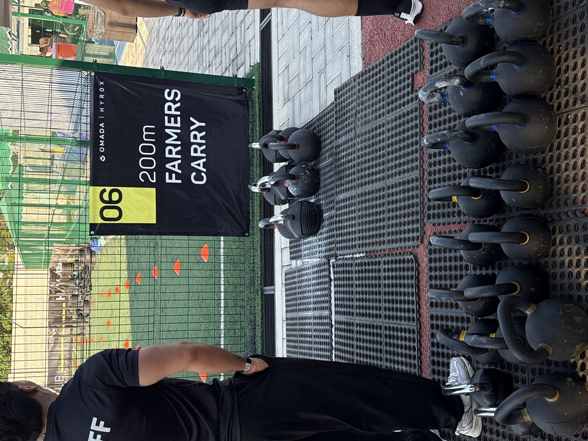 The image shows an outdoor athletic event. The sign reads "200m FARMERS CARRY" under the OMADA | HYROX logo with "06" displayed to the left. To the right are rows of black kettlebells of varying sizes on a rubberized surface. A chain-link fence is visible with a green field and orange cones behind the sign. Two people are partially in the shot, one appearing to be staff, wearing a black shirt with white text, and another in athletic wear. The location is Santa Clara Ocoyucan, Mexico.