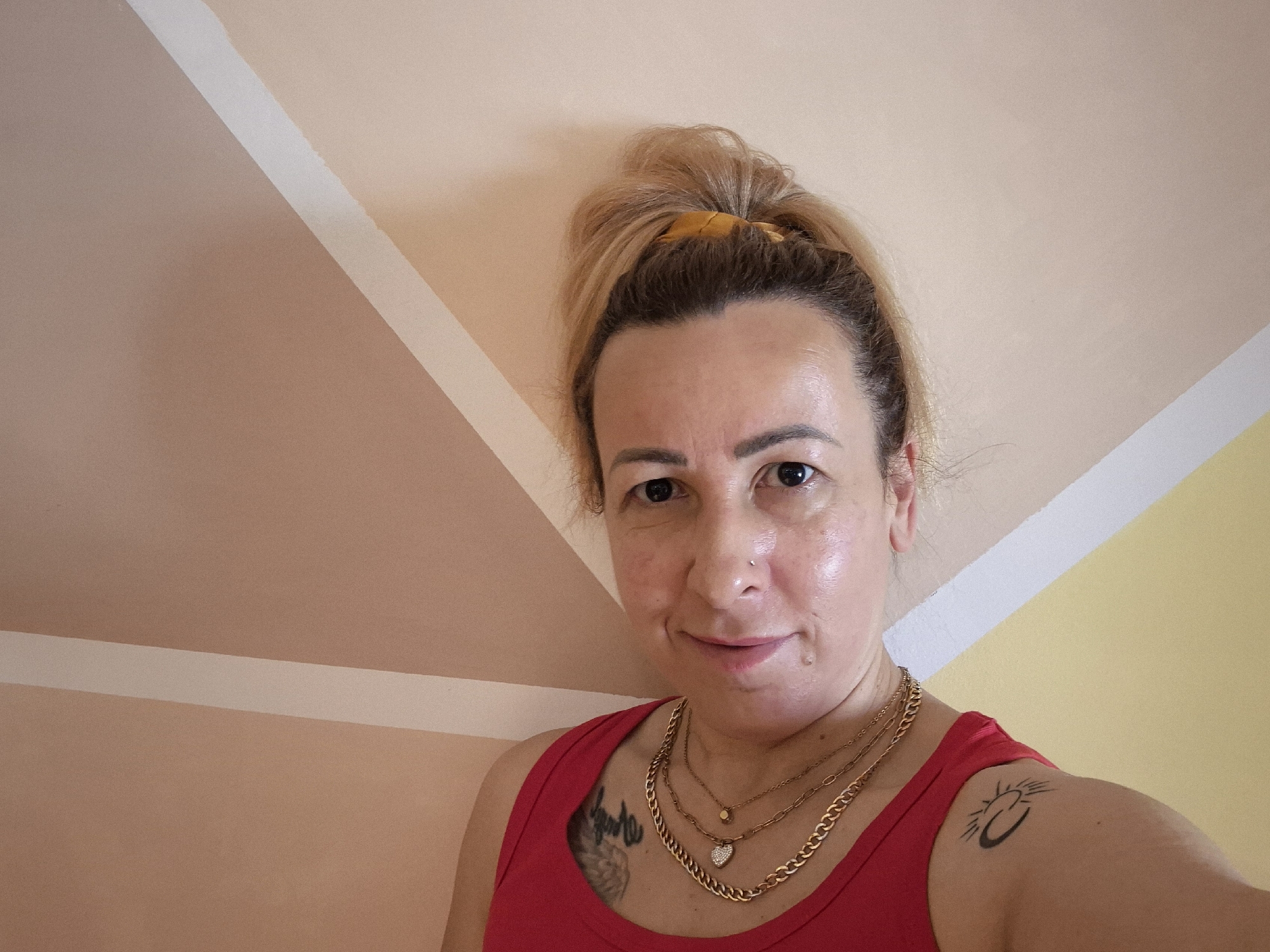 A woman with blonde hair, pulled up in a messy bun with a yellow scrunchie, is captured in a selfie-style shot. She is looking directly at the camera with a slight smile. She wears a red tank top and layered gold necklaces, one featuring a small heart charm with rhinestones. Her skin appears to have a natural sheen, and she has a small nose piercing. Two tattoos are visible: one on her left chest/shoulder area which appears to be a black abstract design or lettering, and another on her right upper arm depicting a stylized black sun and crescent moon design. The background is an interior wall painted with a geometric pattern, featuring sections of light beige, a darker reddish-brown, a bright yellow, and diagonal white stripes separating these color blocks. The lighting suggests an indoor setting, possibly during the daytime. No text is visible in the image.