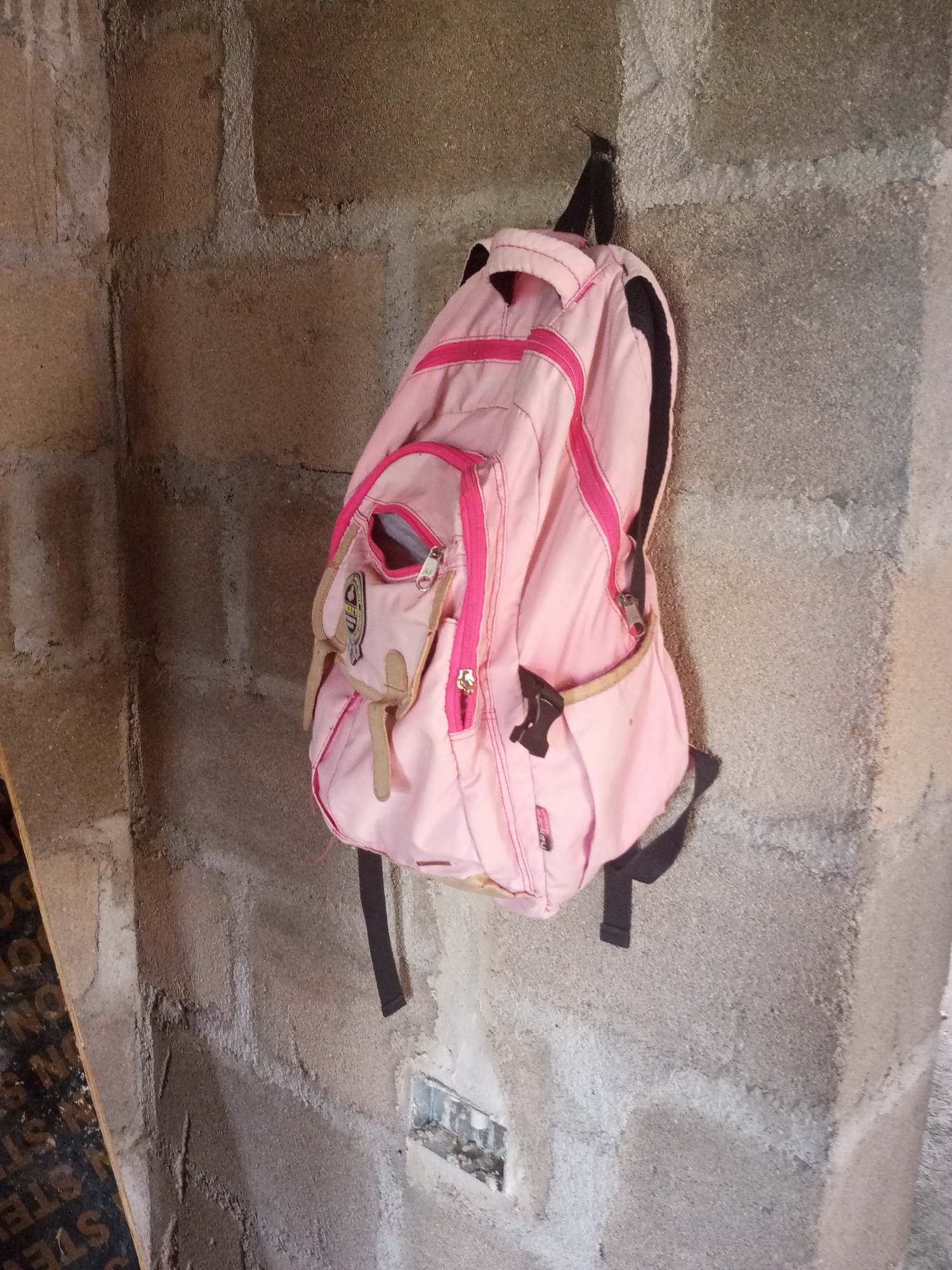 A pink backpack with black and pink trim hangs from a hook on a rough, unfinished concrete wall. The backpack has multiple zippered compartments and pockets. The wall appears to be part of a construction site, with visible rough texture and some unfinished areas. To the left of the backpack, a piece of dark plywood with white text is partially visible. The lighting suggests it is daytime. The image provides no context for the presence of people or any specific activity. There is no visible text on the backpack.