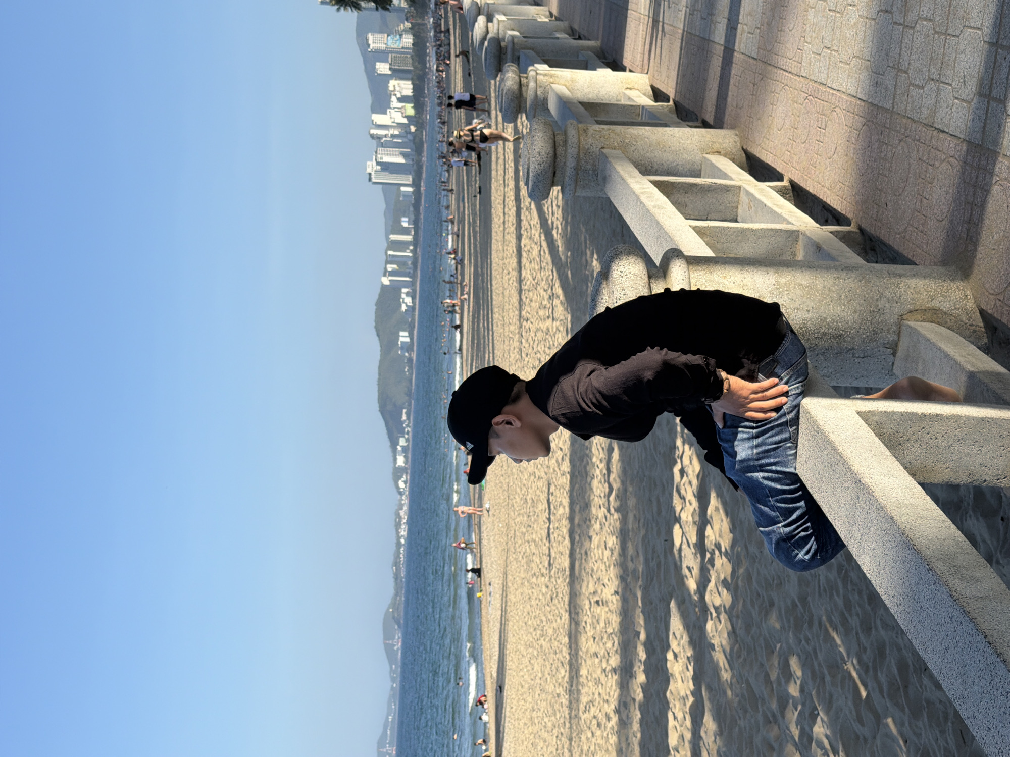 The image captures a young person wearing a black baseball cap and a black long-sleeved shirt, with blue jeans, sitting on a stone barrier overlooking a crowded beach in Nha Trang, Vietnam. The person is facing to the left, looking out at the sea. The beach is sandy, with numerous people scattered across it, some walking, others sunbathing. The ocean is visible, with gentle waves. In the background, a cityscape with tall buildings and mountains is discernible, characteristic of Nha Trang's coastal urban landscape. The sun is casting long shadows across the sand and the walkway. The stone barrier the person is sitting on appears to be part of a promenade or seawall.