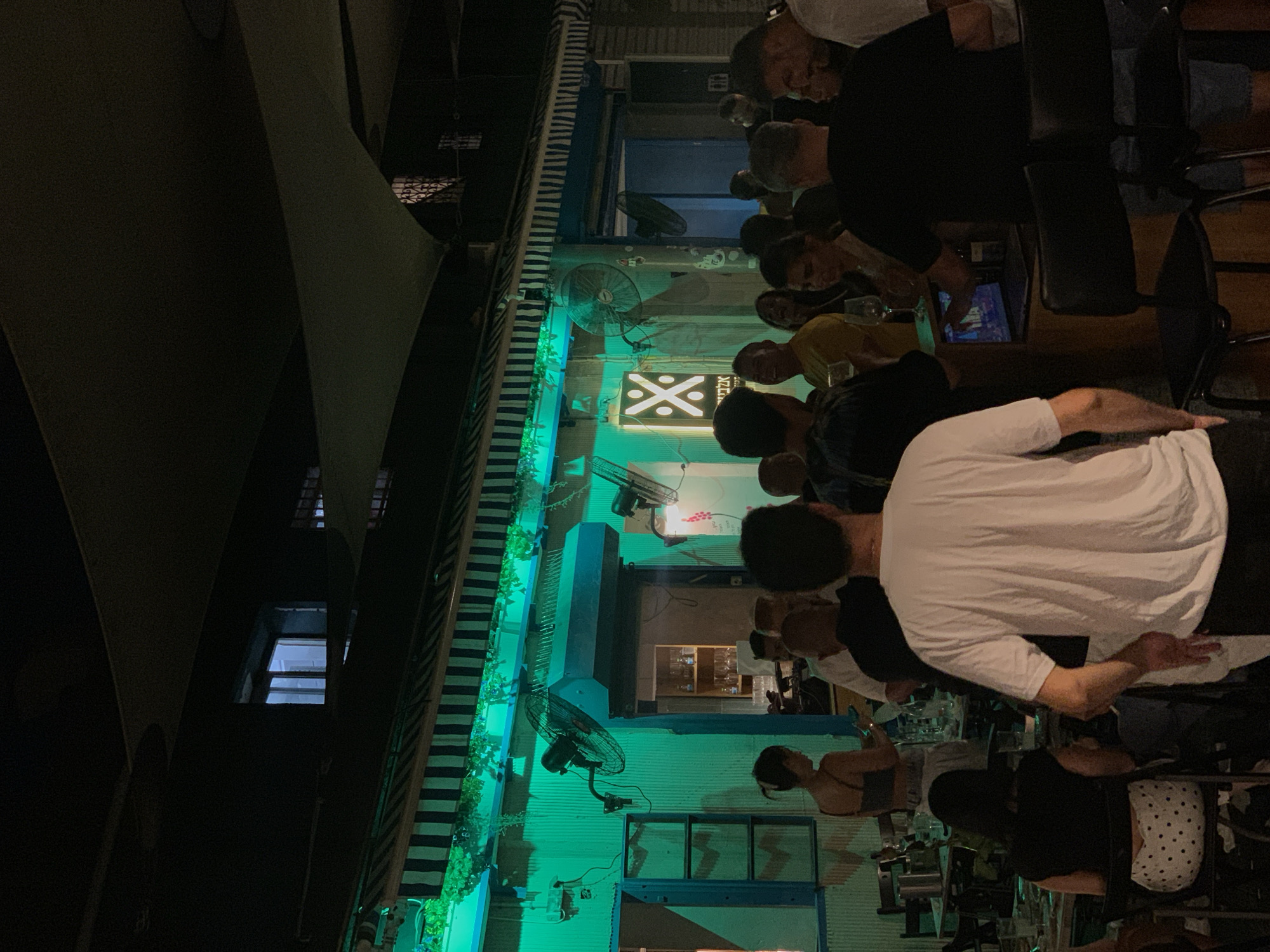 The image depicts an outdoor seating area of a bar or restaurant at night, illuminated by bright green lights from the facade of the building. The establishment appears to be located in Tel Aviv, Israel, based on the Hebrew text visible on a sign featuring a white geometric logo. Several groups of people are standing and seated, engaged in conversations and socializing. Some individuals are holding drinks. In the background, there's a blue-framed doorway leading into the establishment, and several industrial fans are mounted on the wall. Tables with chairs are set up in the foreground and midground, with some appearing to be occupied and others set for service. The overall atmosphere suggests a lively evening social gathering.