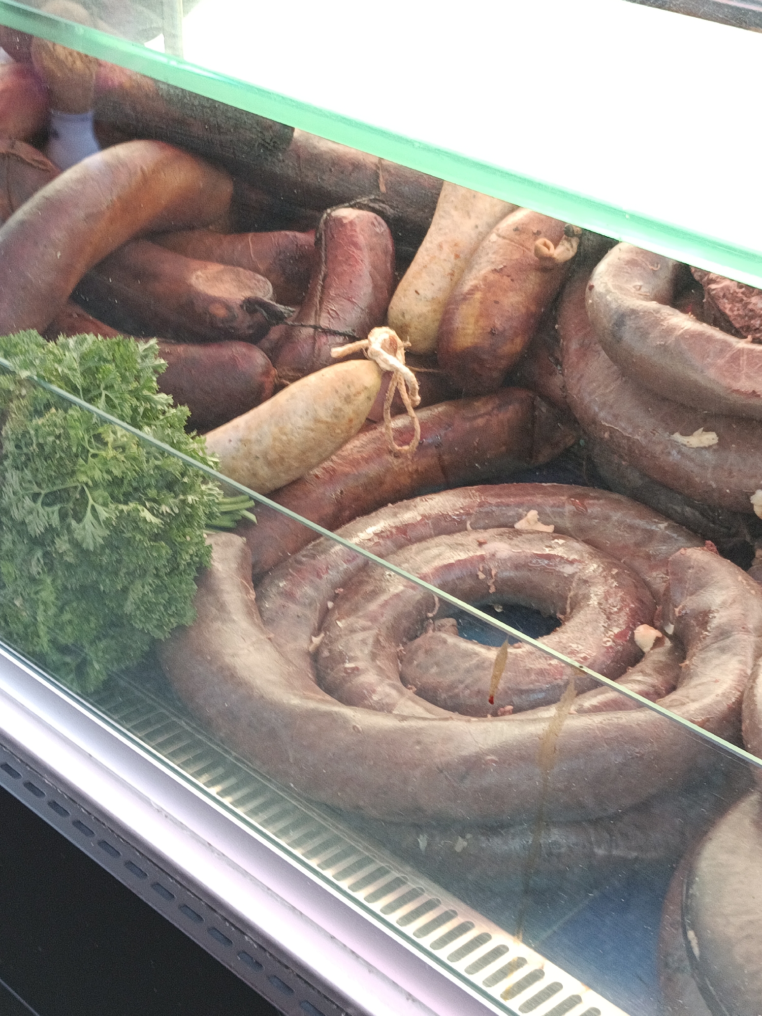The image shows a display of various sausages and what appears to be a bunch of parsley, arranged in a refrigerated glass case. The sausages are of different sizes and colors, some coiled in circles, others in straight lengths. The parsley is a vibrant green and is placed in the foreground to the left. The overall impression is of a food display, likely in a butcher shop or market. The image does not provide any information that allows for determining the location in the city of Le Neubourg, France.