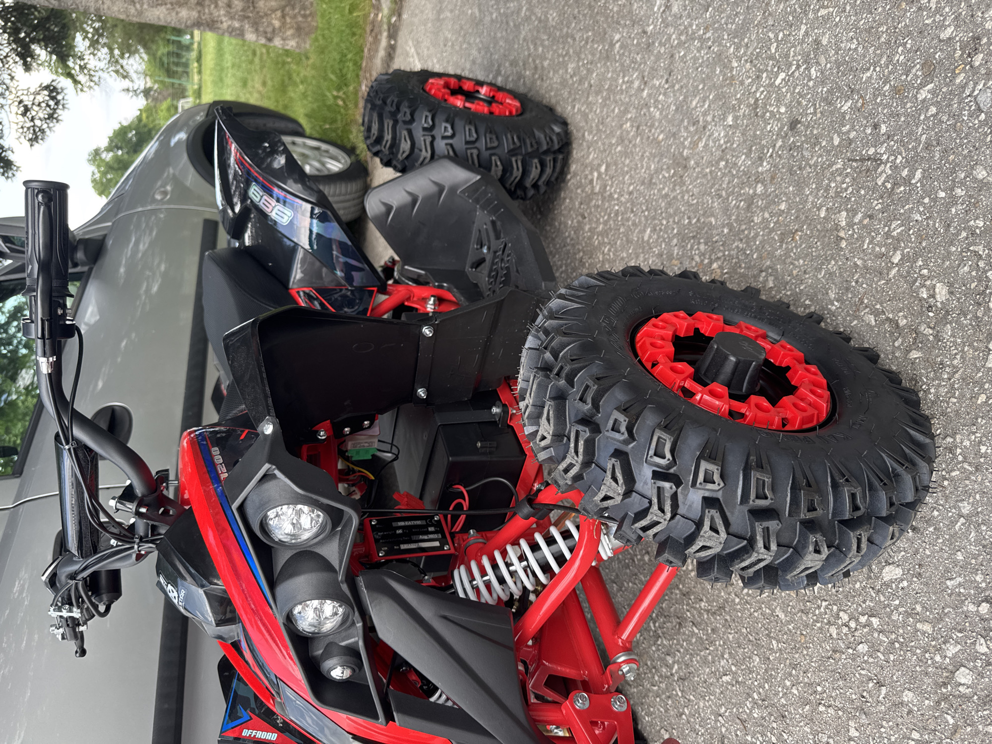 The image captures a red and black all-terrain vehicle (ATV) parked on an asphalt surface. The ATV has large, knobby tires with red rims, indicative of off-road capabilities. The front of the ATV features headlights, and there is a license plate or identification tag visible. Adjacent to the ATV, a gray or silver vehicle is parked, with its wheels partially visible. The background shows a blurred green area, possibly a grassy field or park, and a tree can be seen in the distance. The scene appears to be outdoors in a city setting.