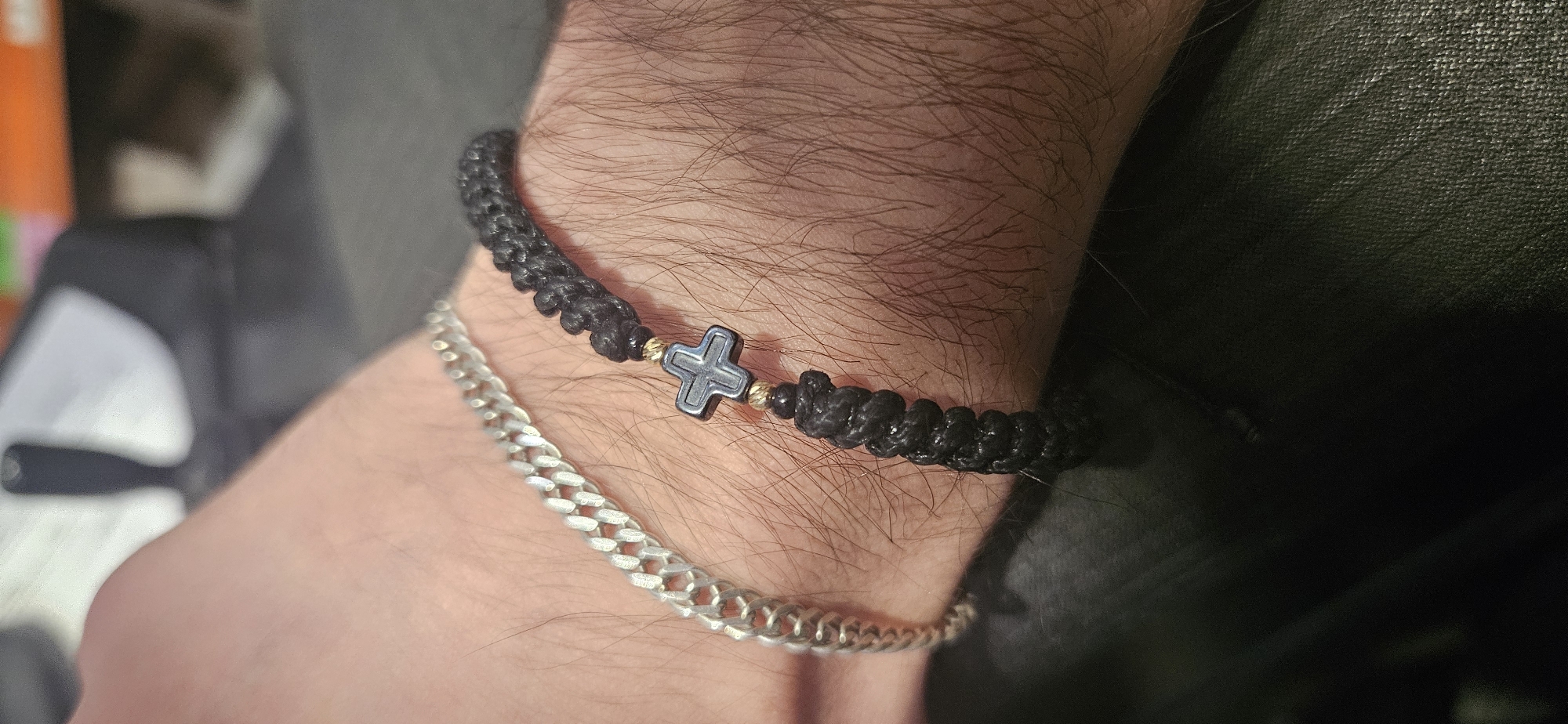 A close-up shot captures a person's hairy forearm and wrist, showcasing two distinct bracelets. The skin tone appears light to medium. The upper bracelet is a black, intricately woven or knotted cord, featuring a dark grey, metallic cross-shaped bead positioned centrally. This cross bead is flanked on both sides by small, gold-colored spherical beads, which appear slightly textured. Below this, a silver-colored chain bracelet, designed in a classic curb chain style, encircles the wrist. The chain links are visible and reflect some light.
The focus is sharply on the wrist and its adornments, while the background is softly blurred and dark, suggesting an indoor setting. Hints of dark, possibly textile material are visible in the background, along with indistinct lighter shapes that are out of focus. No specific activity is depicted beyond the wearing of the jewelry. The lighting suggests an artificial indoor light source, creating subtle shadows and highlights on the skin and jewelry. There is no visible text in the image. While the provided context states the location as Plovdiv, Bulgaria, this information is not visually discernable within the media file itself.