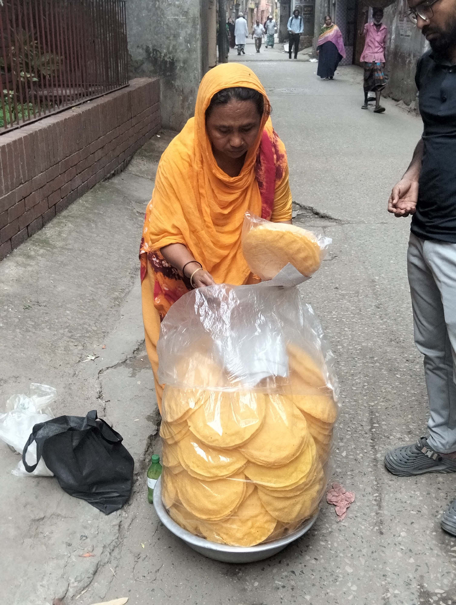 In a narrow street in Dhaka, Bangladesh, a woman in a yellow sari sells papadums. She holds a packaged stack of the thin, crispy wafers, with a large plastic bag full of them in a metal bowl at her feet. A black tote bag and a water bottle sit on the ground nearby. A man stands next to her, appearing to be a customer. Several other people walk in the background of the street, and buildings line the road.