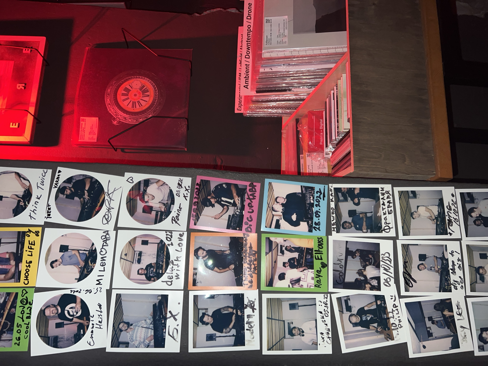 The image shows a wall covered with polaroid photographs and some vinyl records displayed on shelves. The photographs are of various people, mostly men, who appear to be DJs or musicians, often pictured with DJ equipment. The wall appears to be in a dimly lit venue, possibly a club or a music studio. The overall aesthetic suggests a candid and informal documentation of events or performances. The bottom right corner of the image reveals a shelf filled with vinyl records, with labels indicating genres such as "Experimental / IDM / Lo-fi / Abstract" and "Ambient / Downtempo / Drone". One of the record sleeves displays "Aufiassen Aufiassen". The presence of Cyrillic script on some of the polaroids, such as "Емаил Хейстър" and "СМI ЛенОтава", indicates a connection to Bulgaria, and the city of Sofia is mentioned in the metadata as the location. The red lighting in the upper right portion of the image, along with the vinyl records, suggests a possible music venue or record store.