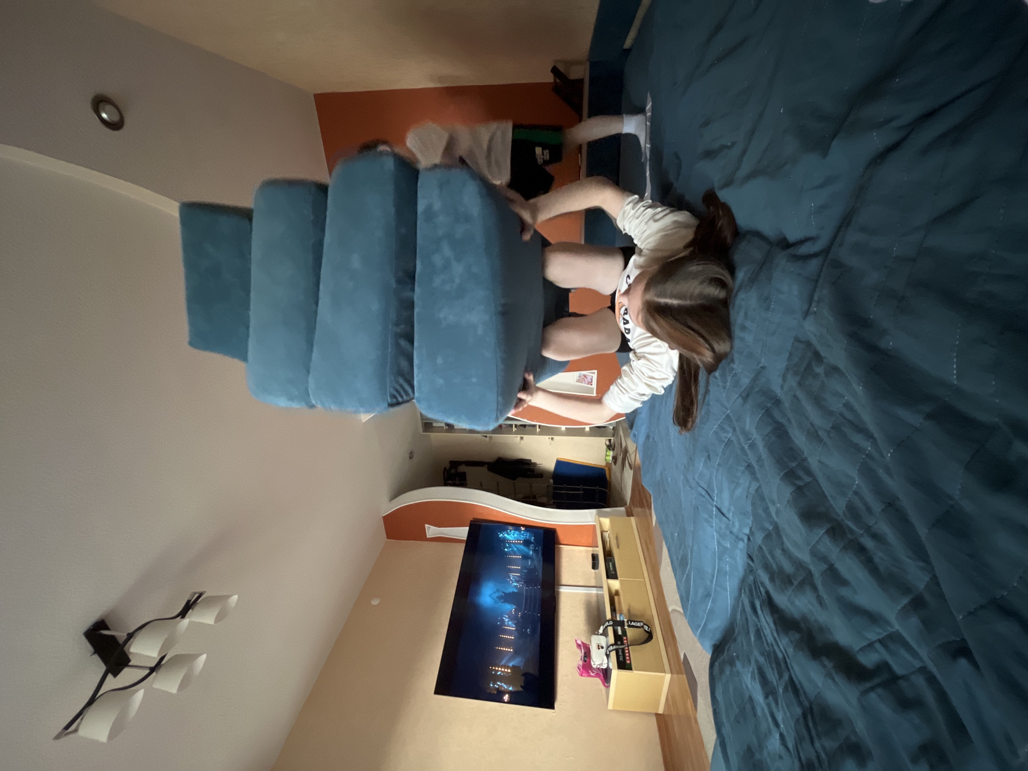 A young woman is kneeling on a large blue bed in a room with light orange walls and a TV set. The room is in Chaikovsky, Russia. A stack of blue upholstered steps sits at the edge of the bed. The woman is wearing a white long-sleeved shirt and black shorts. A second person in black shorts is visible behind the steps.