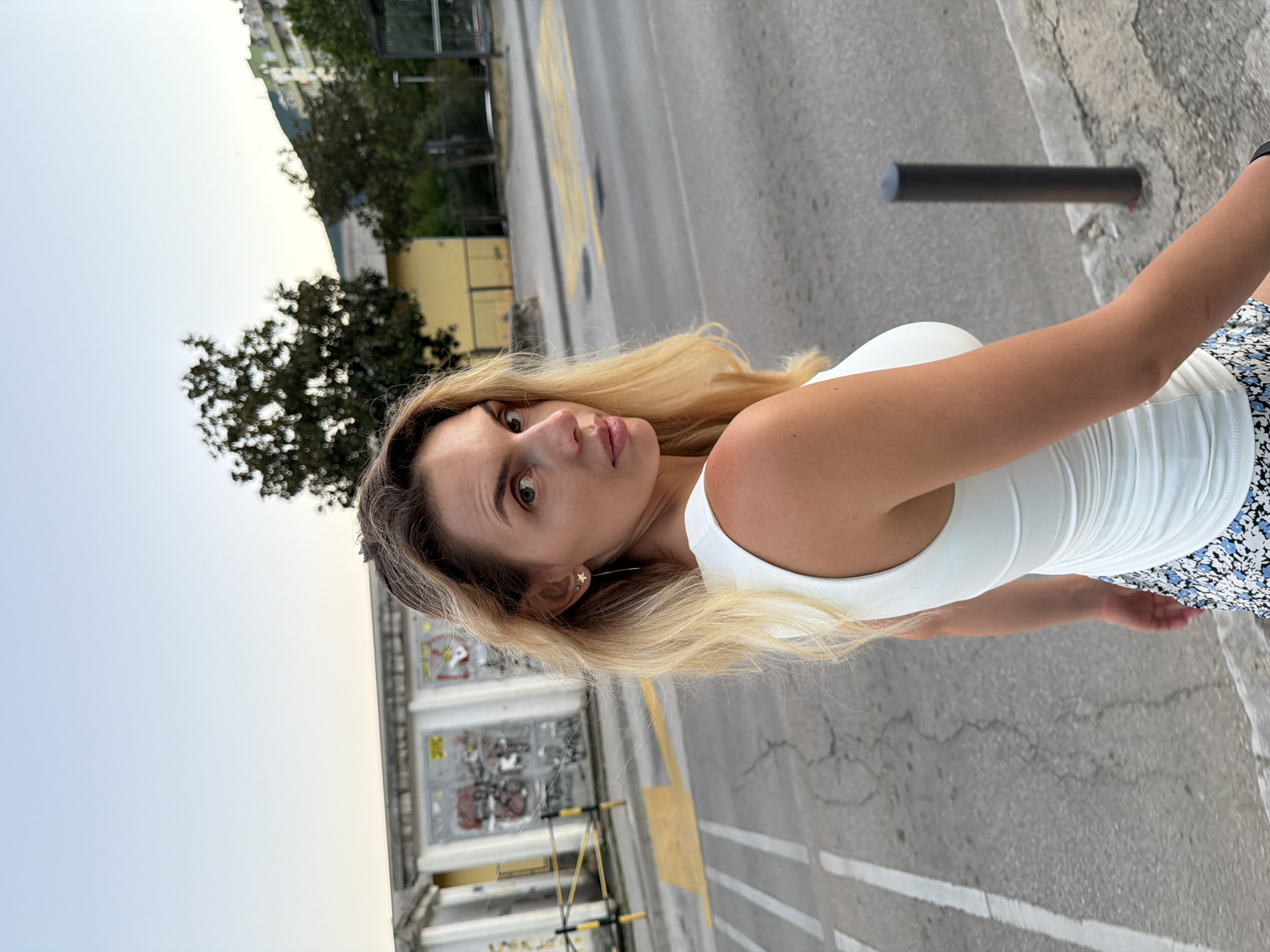 A medium shot captures a fair-skinned woman with long blonde hair and blue eyes, turned away from the camera and looking over her shoulder. She is wearing a white, sleeveless, ruched dress with a floral print skirt. She is standing on a sidewalk, with a street and a graffiti-covered building in the background. The scene is likely Budva, Montenegro, given the architectural style of the building and the surrounding urban environment. The sky is clear and a light blue, suggesting it is daytime, possibly late afternoon due to the soft lighting.