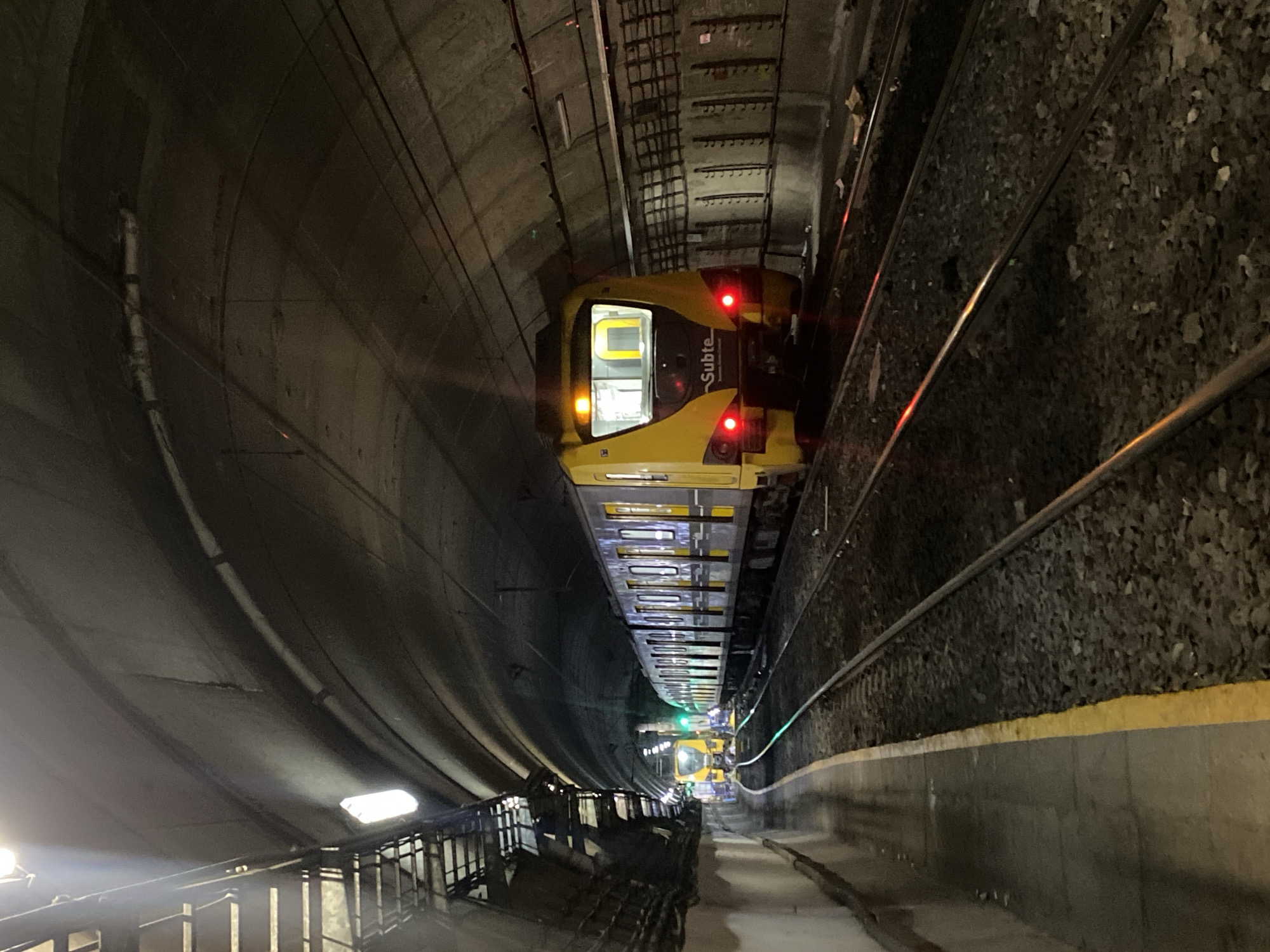 The image shows a subway train in a tunnel, specifically a "Subte" train, which is the subway system of Buenos Aires, Argentina. The train is yellow and brown with the word "Subte" visible on its side. The tunnel walls are concrete and curved, and the tracks are visible on the right side of the image. There are lights in the tunnel ceiling and on the train. The perspective is from slightly above, looking down the tracks into the distance. The tunnel appears to be dimly lit. Some metal structures are visible in the foreground, near the bottom left corner of the image, near the bottom of the tunnel.