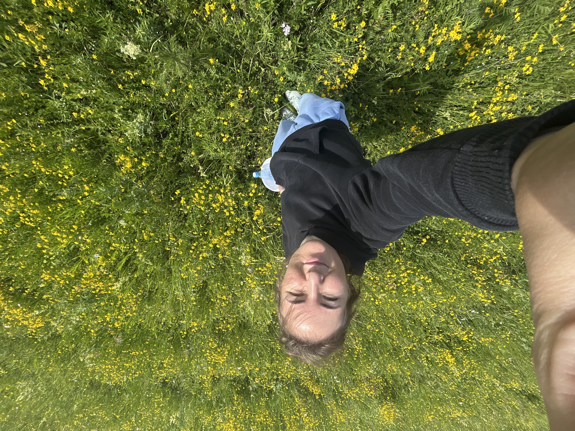 A young woman is lying down in a field of green grass and small yellow flowers. She is wearing a black hooded sweatshirt and light blue pants. A water bottle is visible next to her. The image is taken from a high angle, looking down at the woman and the field. The location is the city of Shabury, Russia.