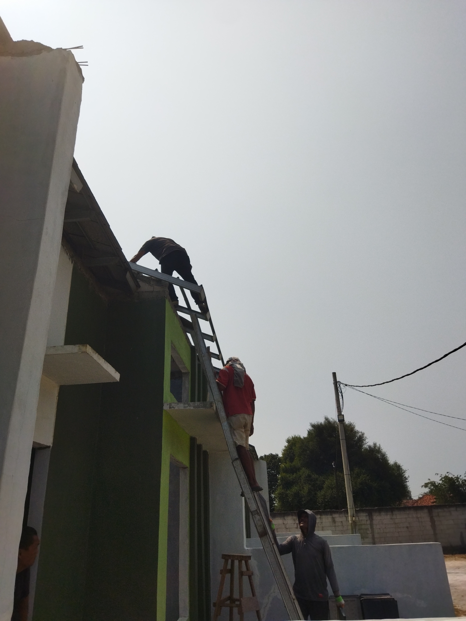 The image depicts an outdoor scene in Suka Sari, Indonesia, where construction is underway. Two individuals are visible on a metal ladder that is leaning against a building. The building has dark green and light green painted sections. One person, wearing a dark shirt and black pants, is positioned higher up the ladder, appearing to be working on the roof's edge. The second person, wearing a red shirt and light-colored shorts, is climbing the ladder below the first.
Further down the ladder, a third individual wearing a grey hooded sweatshirt and dark pants stands at the base, holding onto the ladder with one hand and wearing green gloves. A wooden stool is visible to the right of the ladder, near the base of the building. In the background, there is a brick wall, trees, and a utility pole with electrical wires. The sky is overcast with bright, hazy daylight. The overall impression is of an active construction site on a clear day.