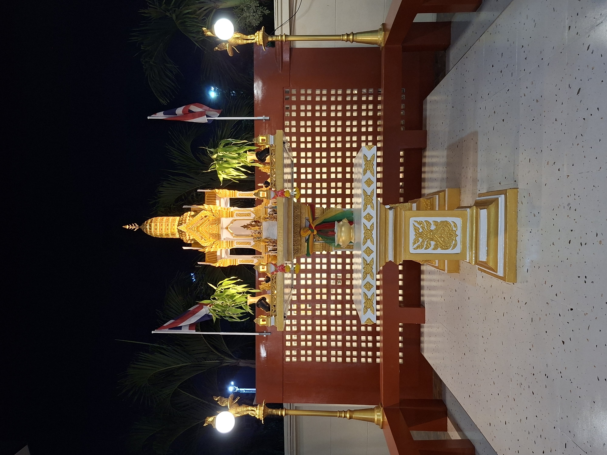 The image shows a spirit house in Pattaya City, Thailand. The spirit house is a small, ornate structure that is believed to be a dwelling place for spirits. It is located on a concrete surface with dark speckles. The spirit house is gold and white, and decorated with Thai flags and greenery. Behind the spirit house is a dark red structure with a grid of square cutouts. Above it, there's a streetlight with a golden figure supporting the bulb. The sky in the background is dark, indicating it is nighttime. Palm trees are visible near the flags.