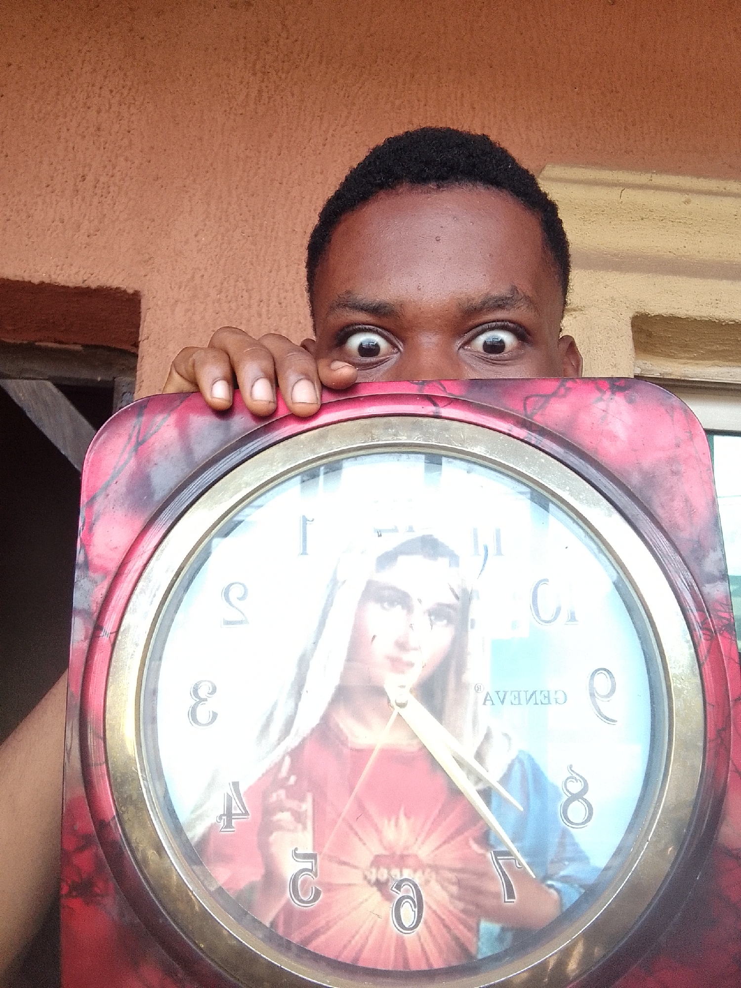 A person with dark skin is holding a large, round, decorative clock in front of their face. Only their eyes and forehead are visible above the clock. The clock face features an image of a person with light skin and a red and blue robe. The clock has a red marbled frame with a gold-colored trim. The background shows a textured orange wall and parts of a window and wooden structure. The location is Nkpor, Nigeria.