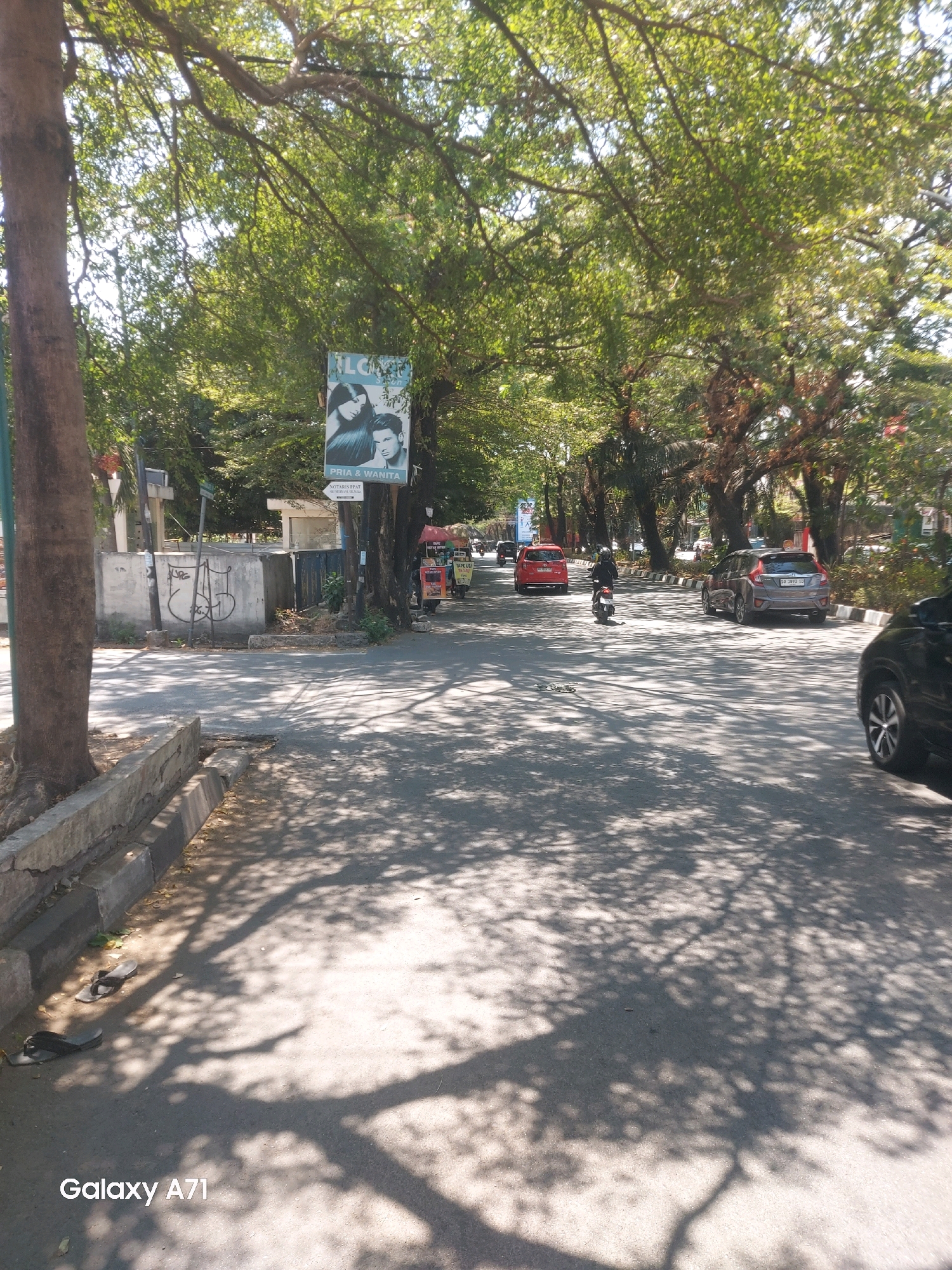 The image depicts a street scene in Makassar, Indonesia, likely during the daytime given the bright sunlight and distinct shadows. A paved road runs through the center of the frame, lined with mature trees that cast dappled shadows across the asphalt. On the left side of the road, a sidewalk is visible, with a tree trunk emerging from it and a concrete barrier. Scattered on the sidewalk are a pair of dark-colored flip-flops.
Several vehicles are present on the road. A red car is moving away from the camera, and a grey hatchback is parked on the right side of the road. A motorcycle with a rider is also on the road, moving in the same direction as the red car. In the background, there are buildings and more trees, suggesting an urban or semi-urban environment. A billboard featuring a man and a woman is attached to a tree on the right side of the road. The overall atmosphere is that of a typical street in a tropical climate. The "Galaxy A71" watermark at the bottom left corner indicates the device used to capture the photograph.