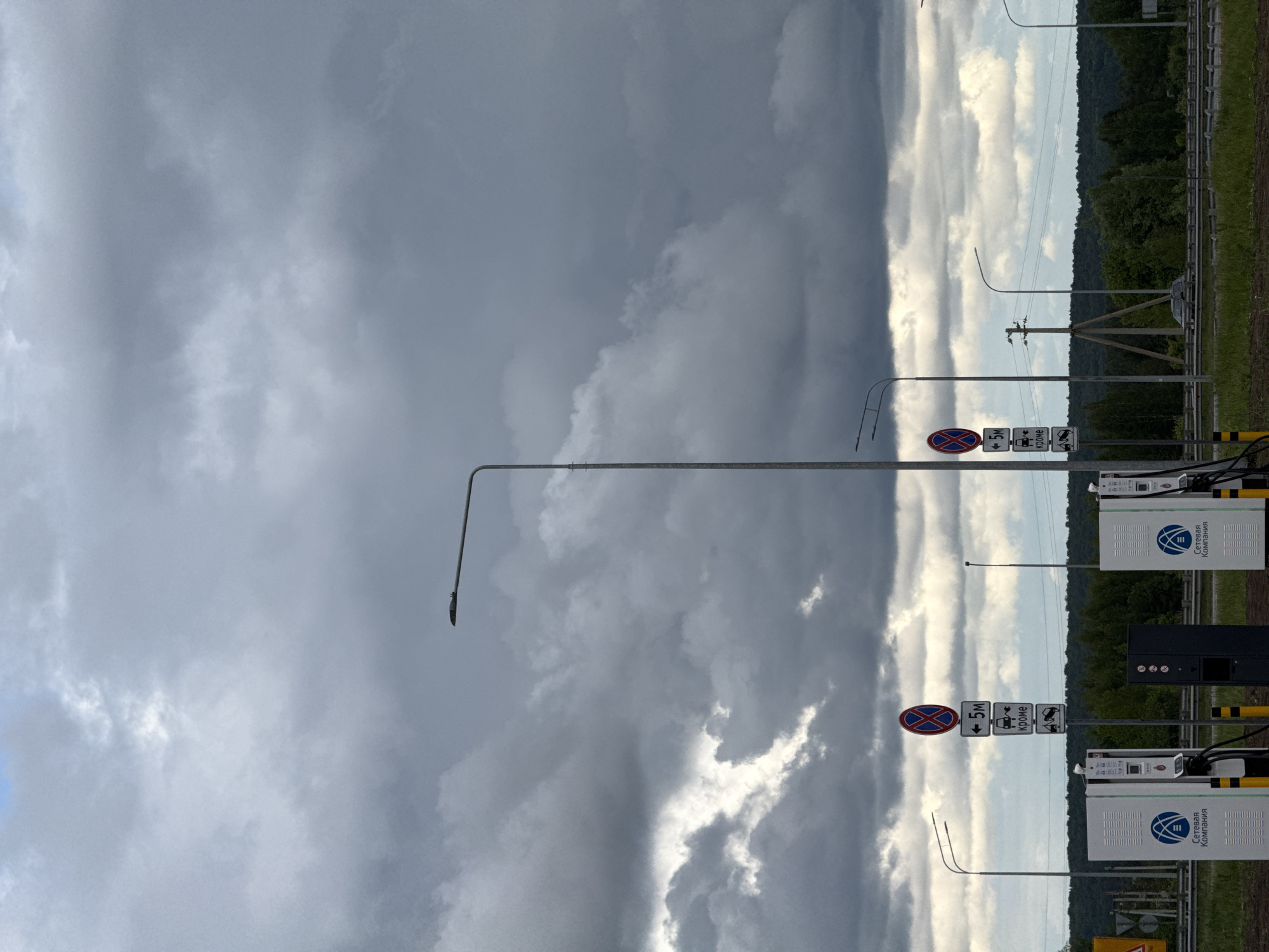 The image shows an outdoor scene in Gil'deevo, Russia, under a dramatic sky with contrasting light and dark clouds. Along a roadside, there are street lamps and regulatory road signs, including a "no stopping" symbol. Two electric vehicle charging stations are visible, branded with "Сетевая Компания." A green strip of vegetation, possibly a forest edge, is present in the background.