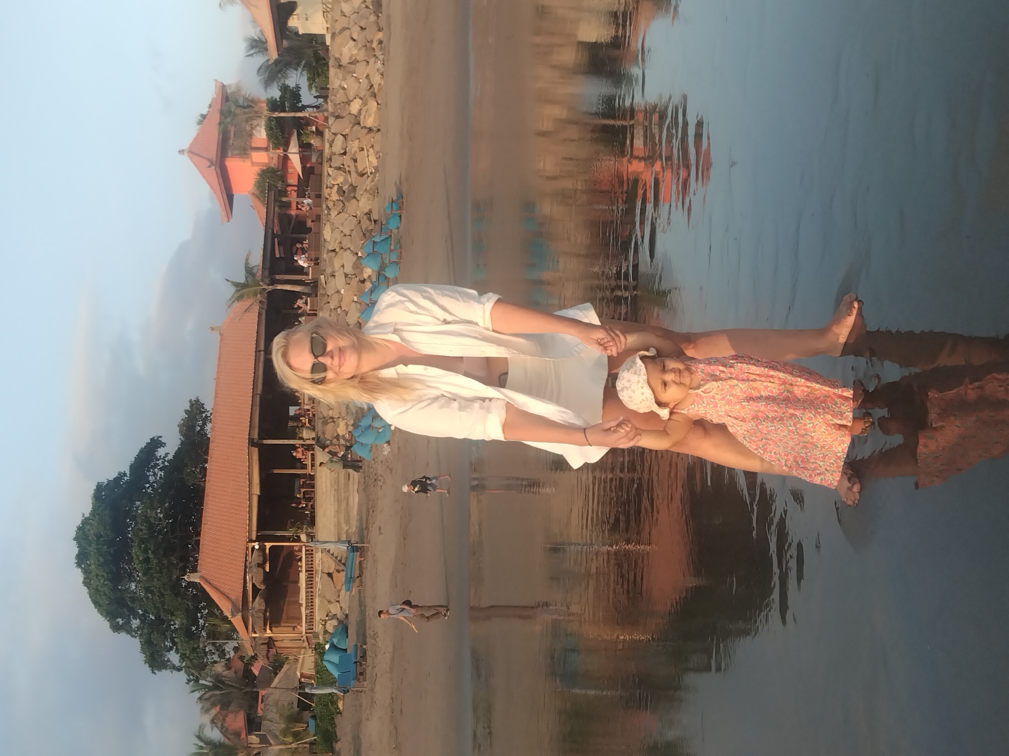 A blonde woman, wearing sunglasses and a white open shirt over a white top and shorts, stands in shallow, still water, holding hands with a young child. The child, dressed in a floral pattern dress and a light-colored hat, is also partially submerged in the water. Both appear to be looking forward, with the woman smiling.
Behind them, the sandy shore stretches back to a series of buildings with distinctive reddish-brown tiled roofs and traditional architecture, reminiscent of Southeast Asian resorts. Lush green trees, including palm trees, dot the landscape between the buildings. Blue beach chairs and small tables are visible along the beachfront, and a stone wall or rocky embankment runs parallel to the water's edge. In the mid-ground, other individuals can be seen walking on the sand.
The time of day appears to be late afternoon or early morning, characterized by soft, warm light and long, clear reflections of the buildings and sky in the calm water. The sky above is a clear, light blue. The overall scene evokes a peaceful and relaxed vacation atmosphere in a tropical setting. No visible text is present.