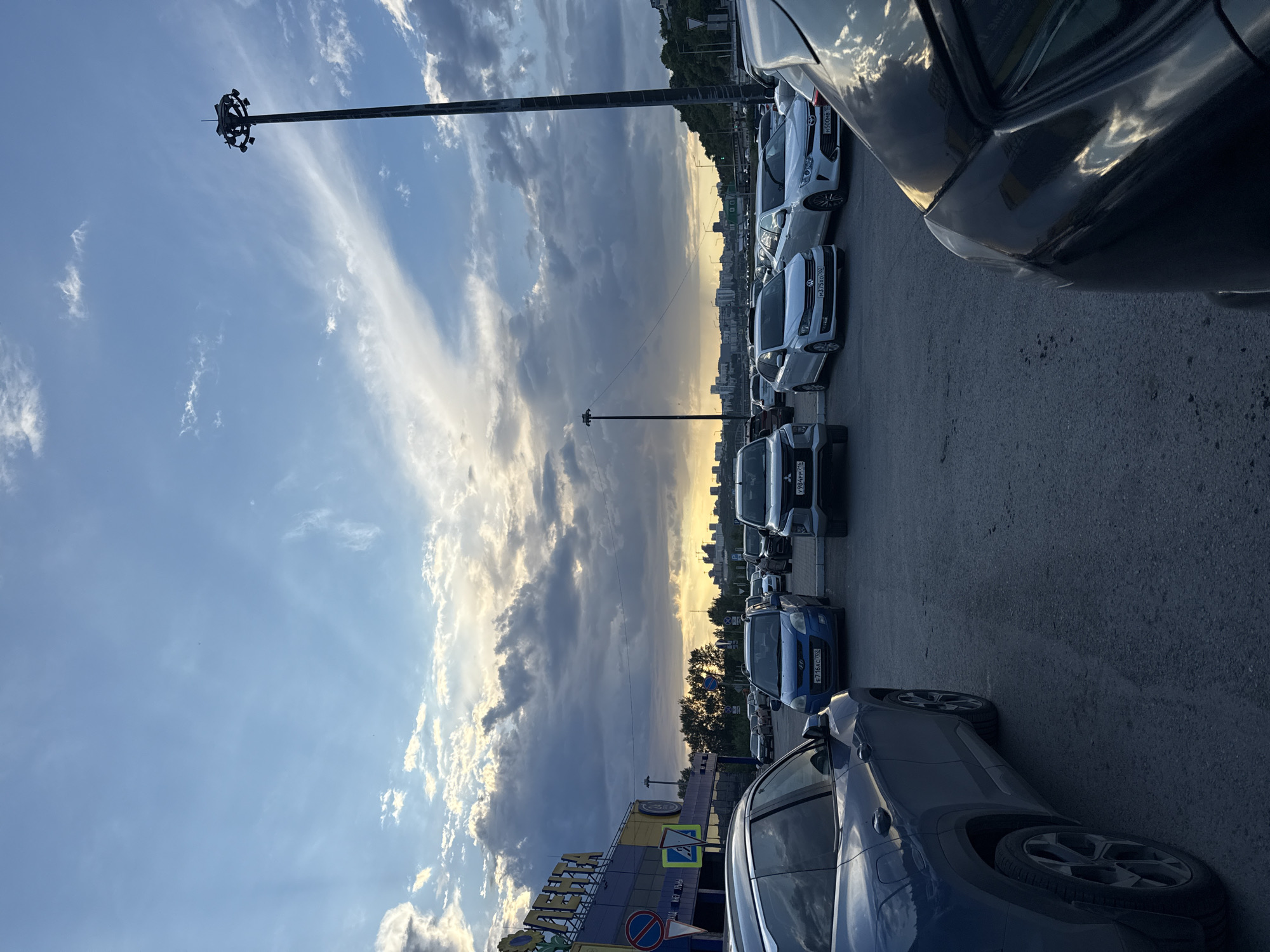 The image depicts a parking lot at dusk, with a mix of cars parked in neat rows. In the background, a city skyline is visible against a dramatic sky filled with clouds and the setting sun. A tall, modern streetlamp stands prominently to the right of the frame, casting a shadow. The foreground features the rear of a dark-colored SUV. A building with signage that includes the word "ЛЕНТА" is visible on the left side of the image. The ground is paved with asphalt, and various traffic signs are present near the building. The overall scene suggests an outdoor commercial or public space during the evening hours.
