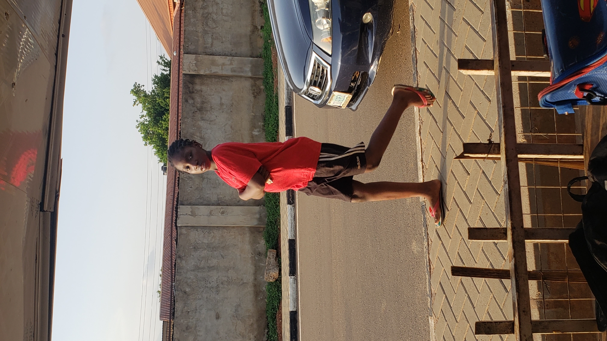 This outdoor image captures a young person standing on a street in what appears to be Narayi, Nigeria. The individual, who has dark skin and braided hair, is wearing a red t-shirt, black shorts with the "ADIDAS" logo on the left leg, and pink and black flip-flops. They are standing with their arms crossed, looking directly at the camera with a neutral expression.
Behind the person, to their left, a dark blue car is partially visible, parked on the paved road. The car's license plate displays "KD 845 J". The road features a curb with black and white stripes, adjacent to a sidewalk made of light-colored, patterned pavers. To the person's right, a metal railing or gate structure is present. A blue backpack with red accents and a cartoon-like character design is visible near the railing, along with a black bag below it.
The background shows a concrete wall or fence, with green trees and parts of building rooftops visible beyond it. The sky is clear and bright, and the long shadows cast by the person and other objects suggest the time of day is either late afternoon or early morning. The overall scene depicts a casual moment on a sunny day in a street environment.
