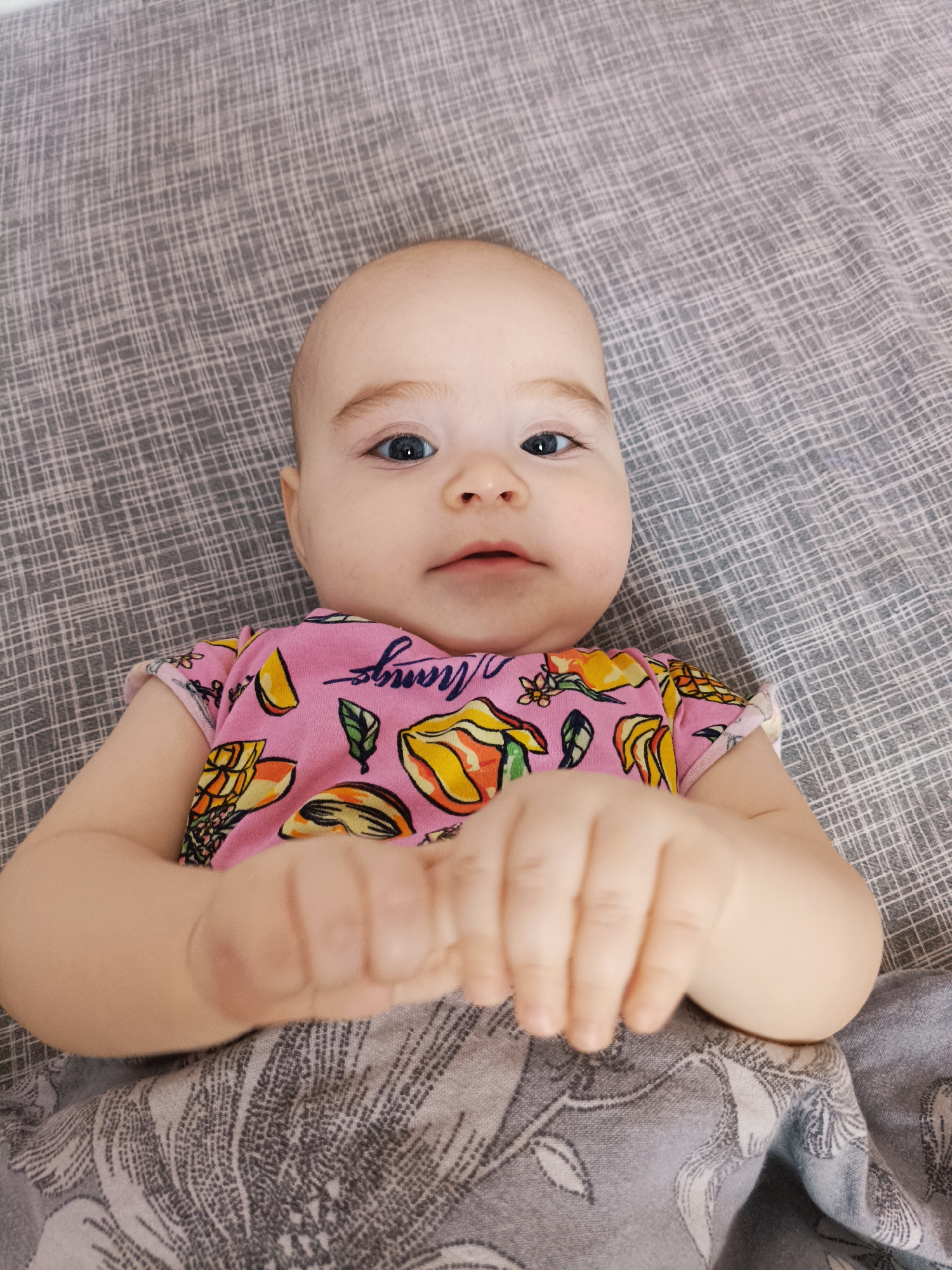 A baby is lying on a gray patterned blanket. The baby is wearing a pink shirt with mango fruit patterns. The location cannot be determined.