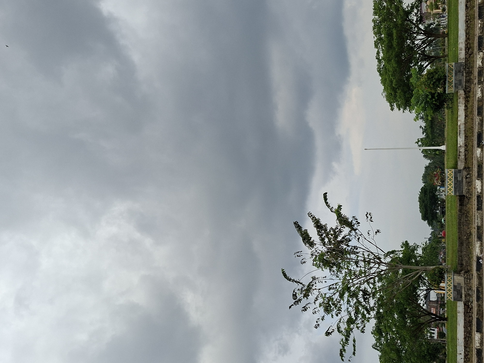 The image depicts a cloudy sky over a park area. A tall, thin flagpole stands prominently in the center, with a lush green lawn and trees lining the perimeter. A small bird is visible in the upper left corner of the sky. A low, decorative barrier or retaining wall runs along the edge of the park, featuring a pattern of black and white markings. The location is Meunasahaluedrienlhoksukon Barat, Indonesia. The environment appears calm and overcast.
