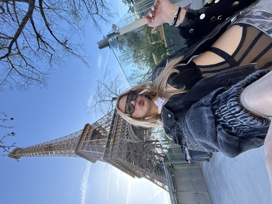 A female is depicted in a close-up selfie, posing outdoors in Paris, France, with the Eiffel Tower as a prominent background element. The subject, with long blond hair and dark rectangular sunglasses, faces the camera with pursed lips. She wears a black denim jacket with embellished shoulders over a white collared shirt and a black, sheer-paneled bodysuit. Her left hand, holding the camera, is visible in the lower left, displaying a silver chain bracelet. Her right hand is raised, showing light-colored nail polish and multiple wristbands, including a red string and a silver-toned bangle.
The Eiffel Tower, a brown iron lattice structure, rises from the mid-left towards the upper center, under a clear blue sky with a faint contrail. To the left of the subject, several black, wheeled trash bins are visible against a metal fence. To her right, a tall, narrow glass panel reflects bare tree branches and the blue sky. Deciduous trees, currently without leaves, frame the upper edges of the image. The ground beneath appears to be a paved surface.