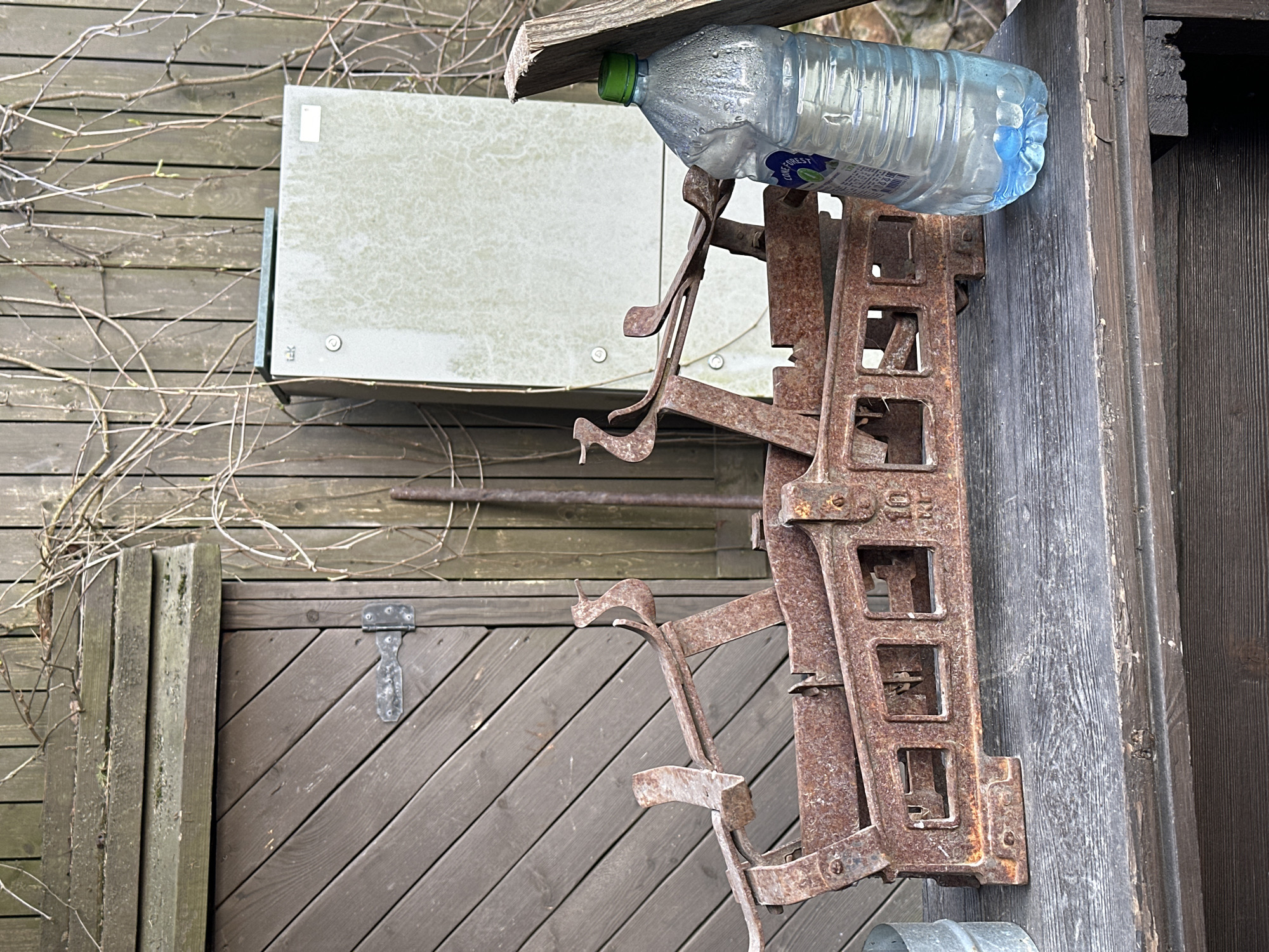 The image shows an outdoor scene in what appears to be a rural or semi-rural area. A rusty, antique-looking metal contraption, possibly a farm implement or a part of a larger machine, is mounted on a weathered wooden wall. A plastic bottle filled with water rests atop the metal structure, partially shaded by a small wooden overhang. The wall itself is constructed from aged, unpainted wooden planks. Adjacent to this wall, a wooden door with a diagonal plank pattern is visible. To the left, a light gray utility box is affixed to another wooden wall covered with bare vines. The overall environment suggests a storage area or the exterior of a building in a rustic or older part of town. There is no visible indication of the location Новоглаголево, Russia.