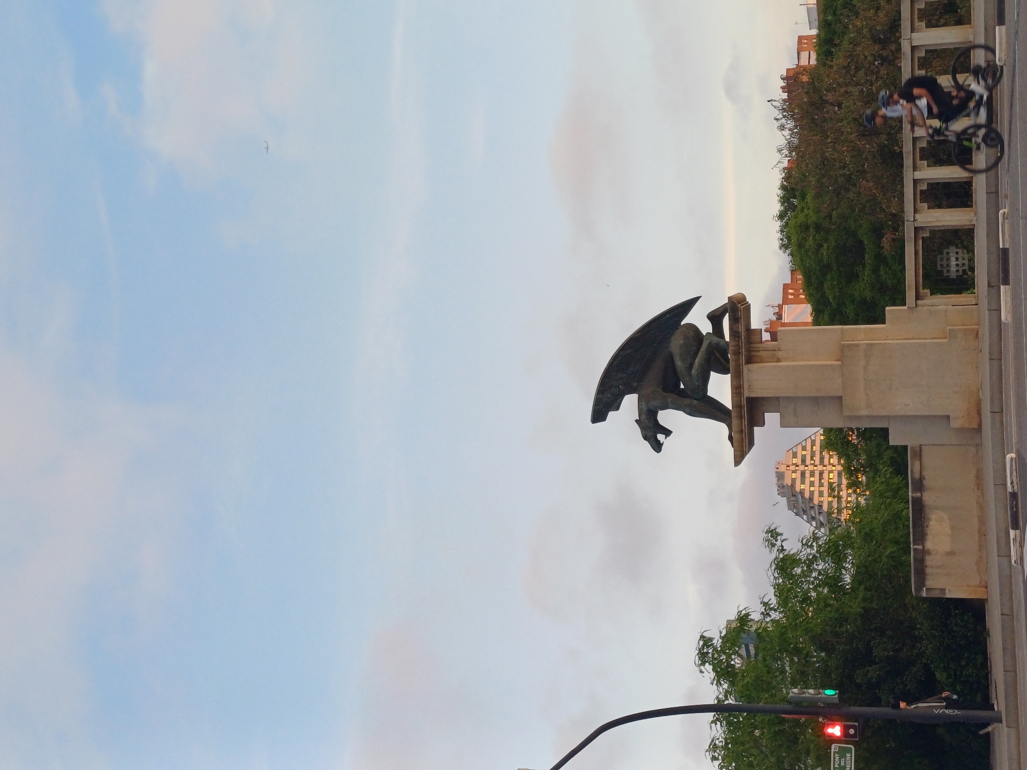 A statue of a winged figure, possibly a gargoyle, is mounted on a concrete pedestal, set against a backdrop of a cloudy sky. A building with a stepped, pyramid-like facade can be glimpsed through the structure. A cyclist is on a bike on the sidewalk. A traffic light pole with the signal is visible. It's located in Valencia, Spain, as indicated by the street sign "PONT DEL REGNE".