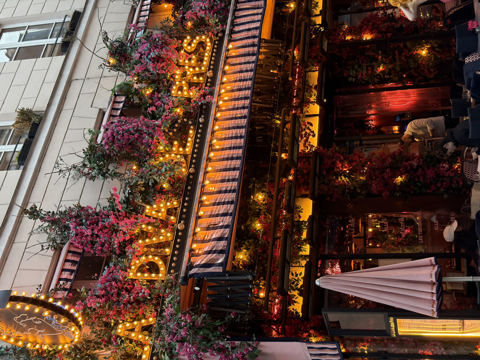 The image captures a vibrant and inviting restaurant or cafe facade at dusk or night, heavily adorned with floral decorations and warm lighting. The original image is rotated 90 degrees counter-clockwise; this description assumes an upright orientation.
On the left side of the frame, a multi-story building with a light-colored, possibly paneled or tiled exterior is visible. Several windows with white frames are present, and beneath some of them, dark rectangular planters hold what appears to be dried grasses or sparse foliage.
The central and right portions of the image showcase the decorated facade of the establishment. A prominent sign features large, illuminated marquee-style letters spelling "SARA..." with the full word obscured but possibly continuing to "SARAH" or similar. Below this, a smaller, circular or oval-shaped sign is also illuminated, but its details and text are difficult to decipher.
Abundant pink and red bougainvillea-like flowers, mixed with green foliage, generously cascade and wrap around the signage and windows, creating a lush, festive look. Numerous small, warm-toned lights are interspersed within the greenery, adding to the glowing atmosphere. A striped retractable awning, featuring dark blue (or black) and light blue (or white) stripes, is partially extended over the entrance or outdoor seating area, also lined with similar warm lights.
Further to the right, behind the awning, are large glass windows or doors offering a glimpse into the establishment's interior, which is bathed in warm red and orange light. Several people are visible inside or near the entrance; one person in a light-colored top (possibly white or light grey) is standing.
In the lower right corner, a light pink or beige parasol (umbrella) is seen in a closed position. Below it, an outdoor heater emits an orange glow, indicating an outdoor seating area.
Visible text includes "SARA..." on the main sign. On the closed parasol, the words "LA CASA DES FRITES" and "BLLEZ-VOU" are partially legible. Through the restaurant windows, snippets of text such as "RESTAURANT," "LES TROUVAILLES," and possibly "BAR" and "BIERE" can be faintly discerned.
The overall impression is one of a lively, cozy, and romantically lit venue, likely a restaurant or bar, during the evening hours, possibly in an urban setting.