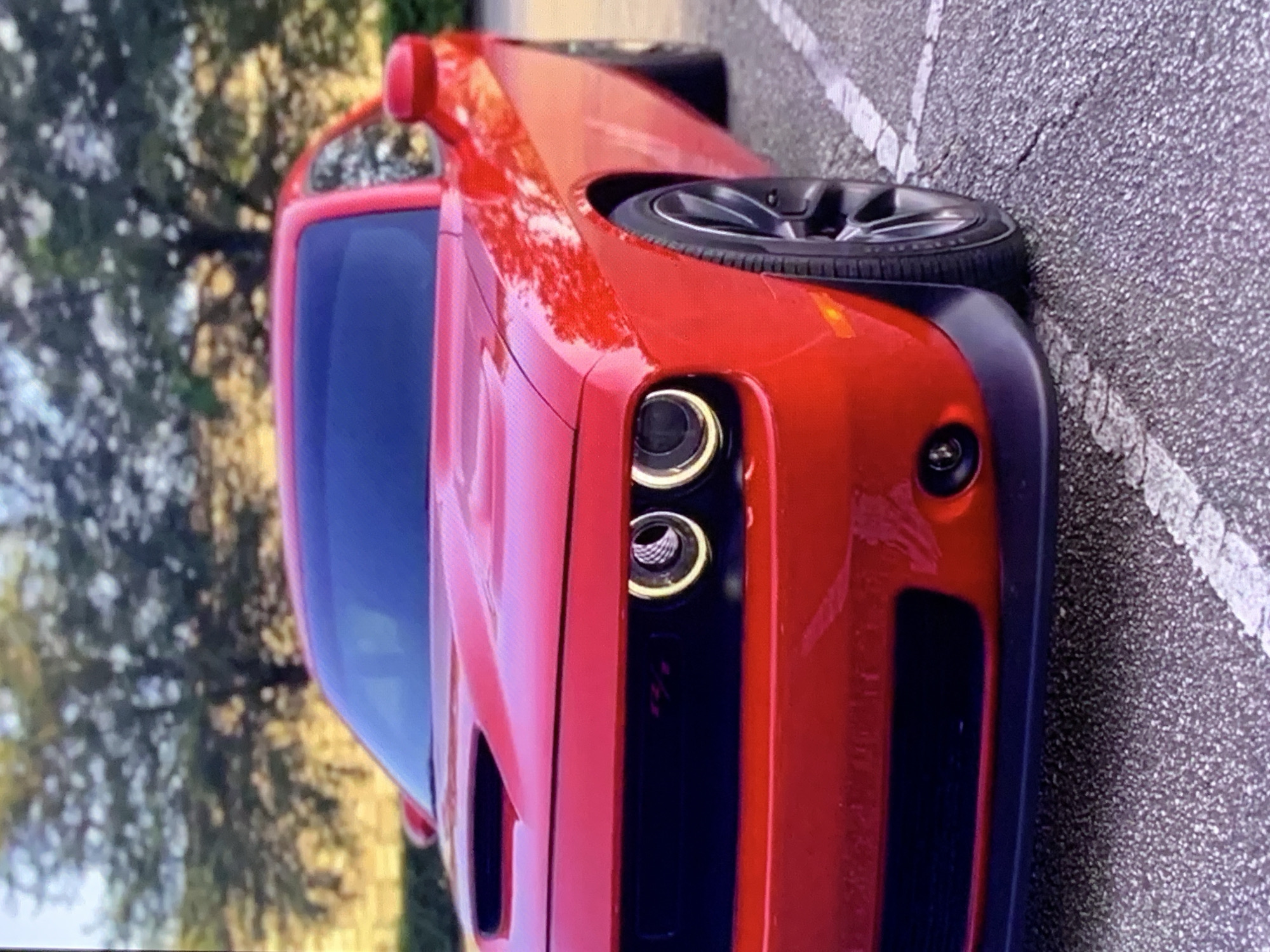 A bright red Dodge Challenger is parked in a parking lot. The car is facing left and is partially obscured by the edge of the frame. The car's headlights are on, illuminating a halo-like ring around the main bulb. The front grille is black and features a red emblem that appears to say "SRT". The car has a hood scoop and a black front splitter. The background is blurred and shows trees and foliage. The parking lot surface is asphalt with white lines indicating parking spaces.