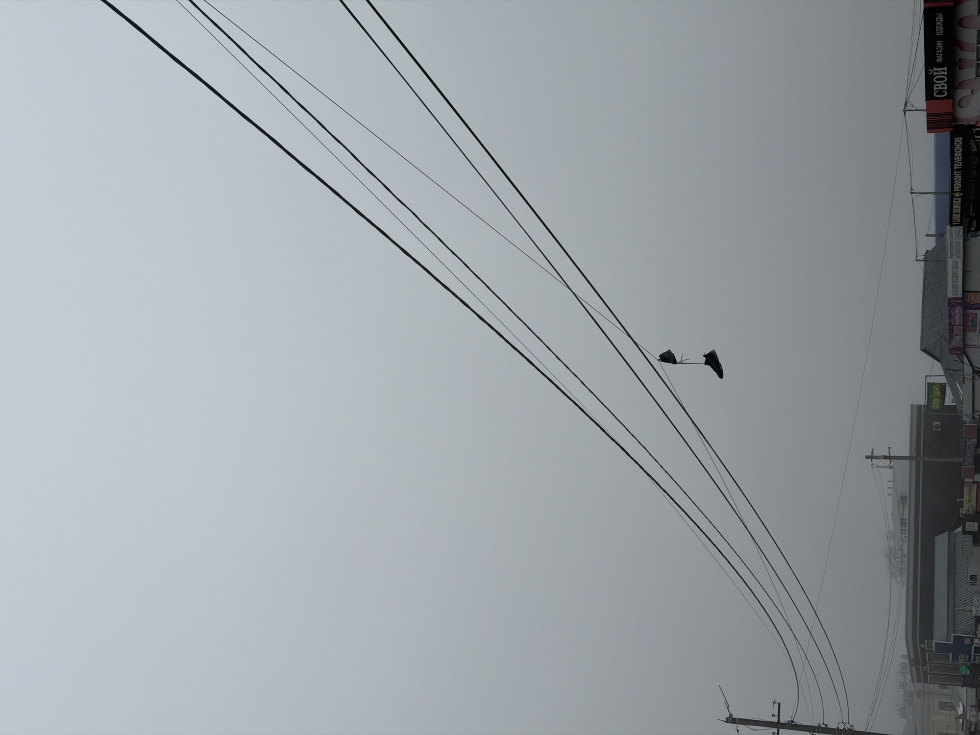 The image, rotated 90 degrees counterclockwise, presents an overcast day in Sarapul, Russia. In the lower right quadrant, buildings are visible, some with commercial signage in Russian script. Overhead, black power lines extend diagonally across the frame, and suspended from them are two shoes. The sky is a uniform gray, indicating heavy cloud cover. Details regarding people or specific actions are not discernible in the image.