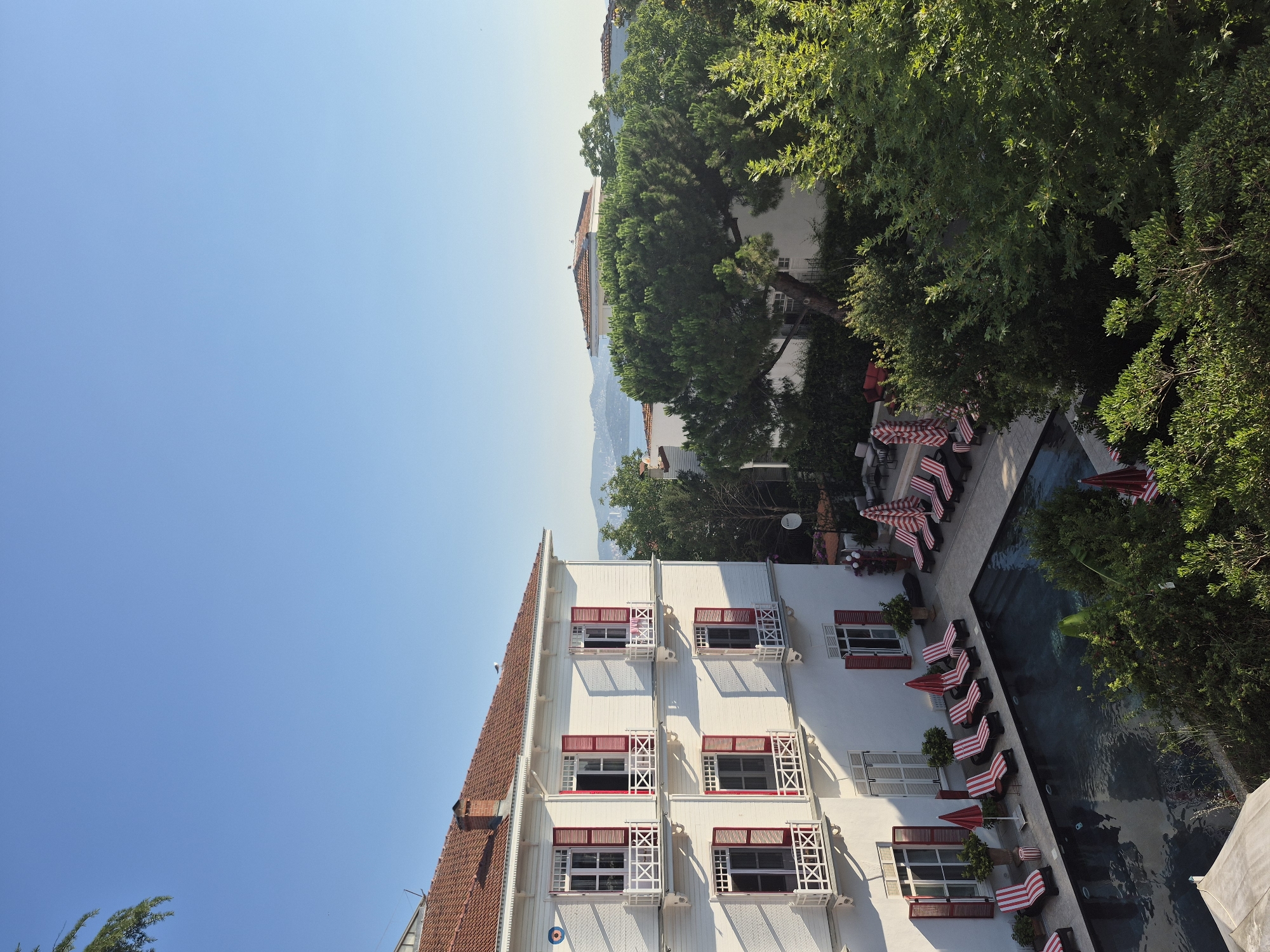 This is a high-angle, full shot of a white, multi-story building with red-shuttered windows and a red-tiled roof, typical of architecture found in the Princes' Islands, Turkey. The building is situated next to a dark, rectangular swimming pool surrounded by a paved area and lined with red and white striped lounge chairs and umbrellas. Lush green trees and foliage are prominent around the pool and building, with more distant trees and rooftops visible. The sky is a clear, bright blue, suggesting a sunny day. There are no people visible in the scene. The overall impression is one of a tranquil, possibly resort-like setting.