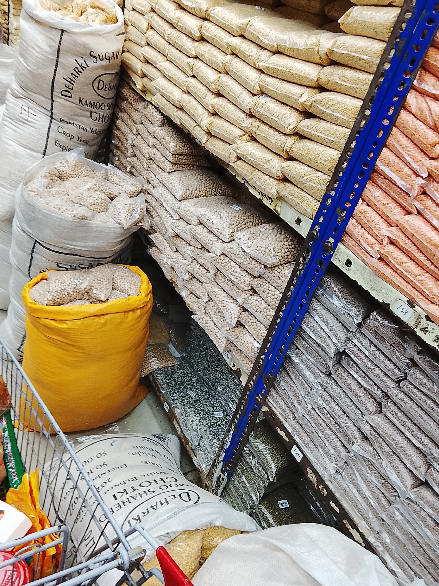 The image shows a well-stocked grocery store shelf filled with various beans, lentils, and sugar in Gulgasht Colony, Pakistan. Large sacks of "Deharki Sugar" and "Deharki Moo Shaheed Ghotki" can be seen. Smaller, packaged quantities of different beans and lentils are arranged in neat rows on the shelves. The store shelves are metal and painted blue. A shopping cart with groceries can be seen in the foreground.
