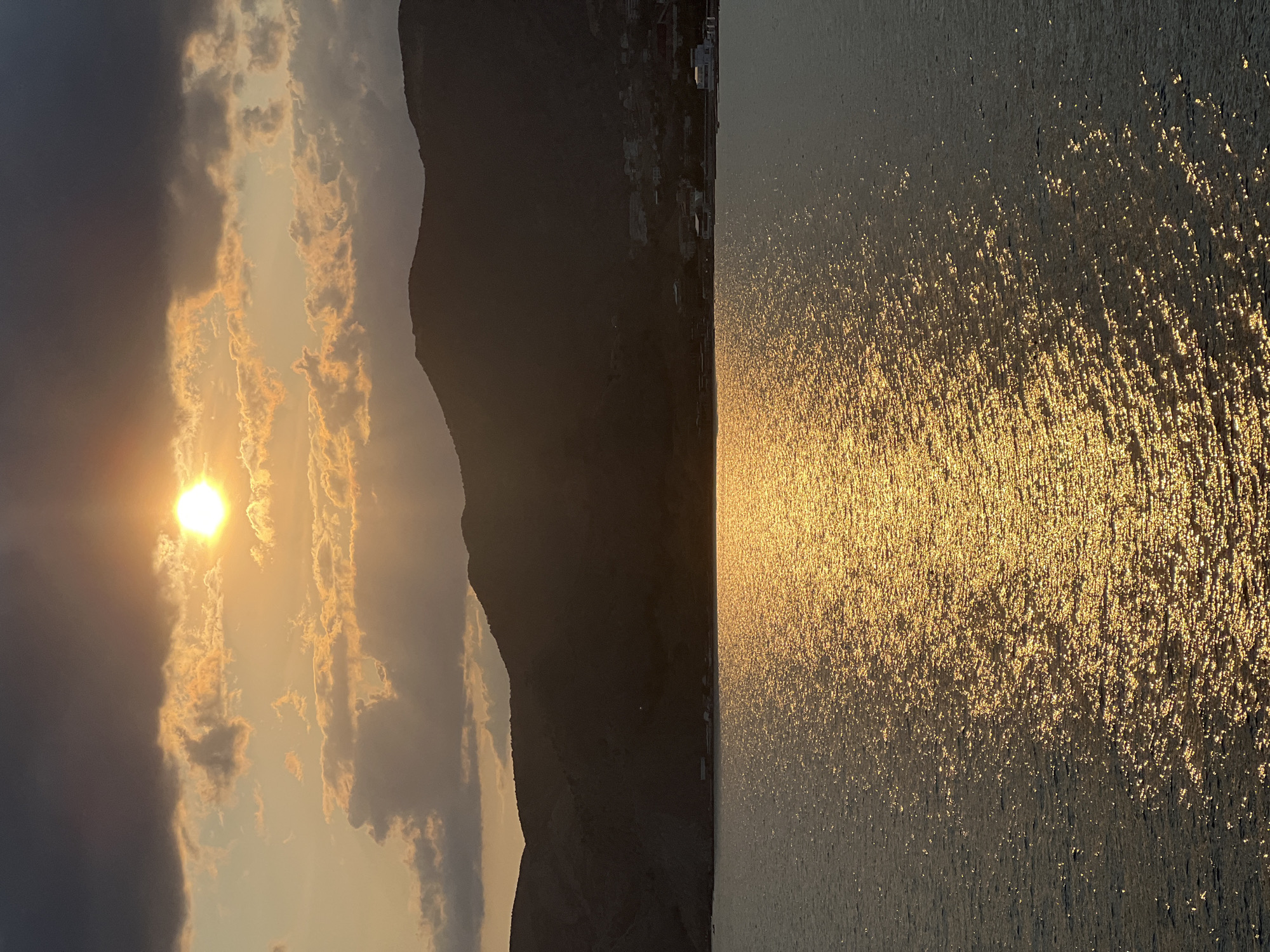 The image depicts a serene coastal scene at sunset in Sudak, Ukraine. The sun, a bright orb, is partially obscured by dramatic clouds, casting a warm, golden glow across the sky and water. The sea reflects the sunlight, creating a shimmering, textured surface. In the background, dark, silhouetted mountains rise, their slopes dotted with hints of buildings, suggesting a coastal settlement. The overall atmosphere is one of tranquility and natural beauty, characteristic of a sunset over the Black Sea.