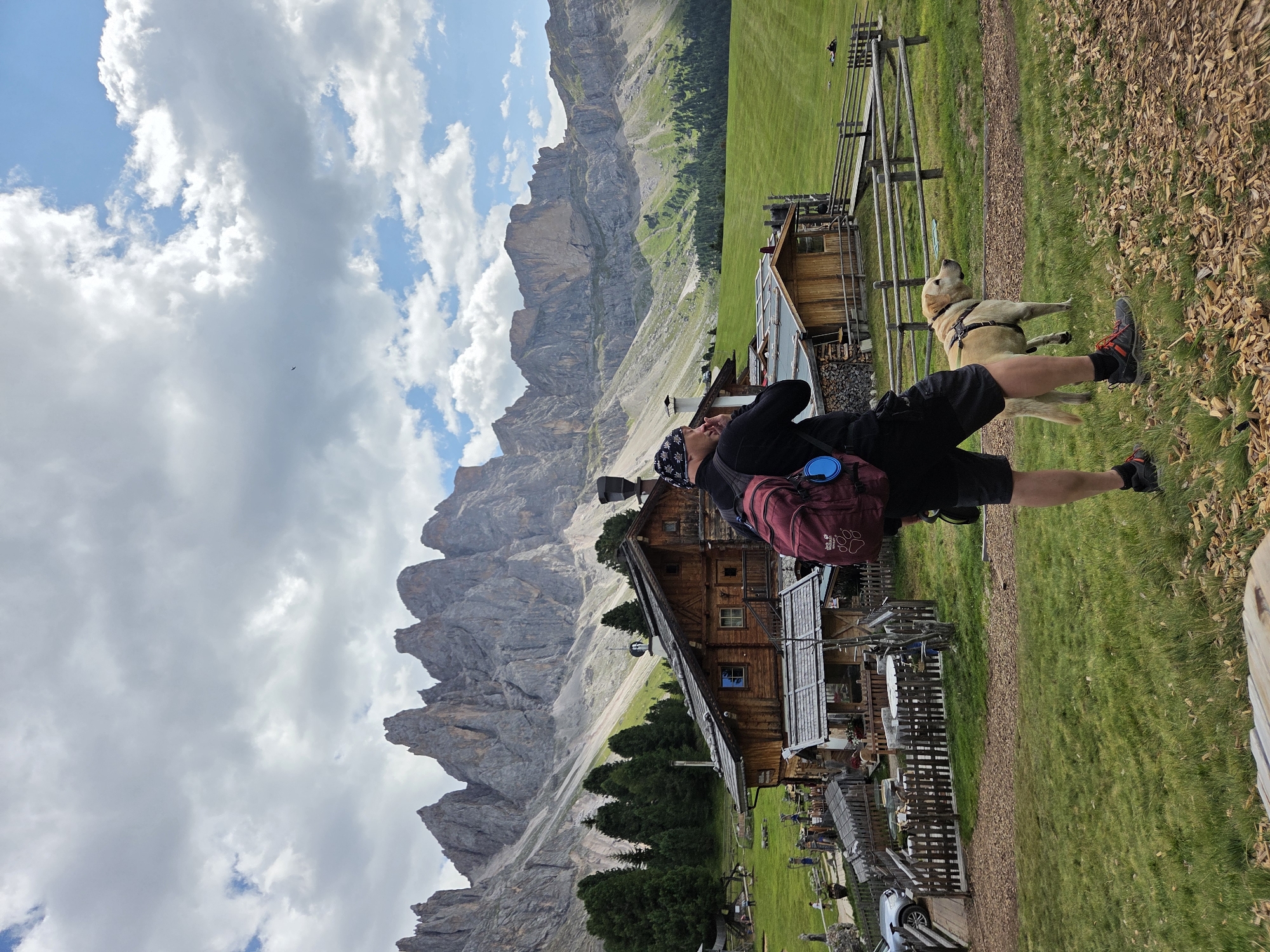 The image shows a person and a dog in a mountainous landscape. The person is wearing a black shirt, black shorts, and a bandana on their head. They are carrying a maroon backpack with a blue water bottle attached. The dog is a light-colored Labrador or similar breed, wearing a harness.
The scene is set in the Italian Alps, with dramatic, jagged mountains in the background. There is a wooden building, likely a mountain hut or lodge, with solar panels on its roof. In front of the building, there are tables and chairs, suggesting it's a place for rest or dining. The ground is covered in lush green grass and some scattered dry leaves. The sky is partly cloudy with a bright blue sky peeking through.
The person appears to be looking up at the mountains, possibly taking a photo or simply enjoying the view. The dog is looking in the same direction. The overall mood seems peaceful and adventurous, typical of a hiking or outdoor excursion in a scenic location. The lighting suggests it is daytime, likely late morning or early afternoon, given the brightness of the sun. No visible text is present in the image.