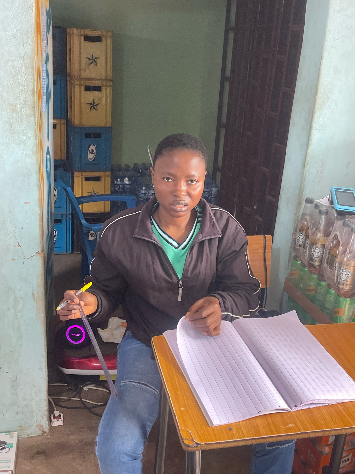 A young Black woman is sitting at a wooden table with an open notebook in front of her, in what appears to be a storage or small shop area. She is wearing a black jacket over a green shirt and blue jeans. She holds a yellow pen in her right hand, and a ruler is positioned between her fingers and the pen. To her left, a red and black appliance with a glowing purple circle is visible, along with electrical cords and a white plug. In the background, there are stacks of blue and yellow crates, some containing bottles. Bottles of beverages are also stacked on a shelf to her right, alongside a small blue electronic device. The environment seems to be an indoor space with a concrete floor and green walls. The scene is likely located in Jos, Nigeria, given the context.